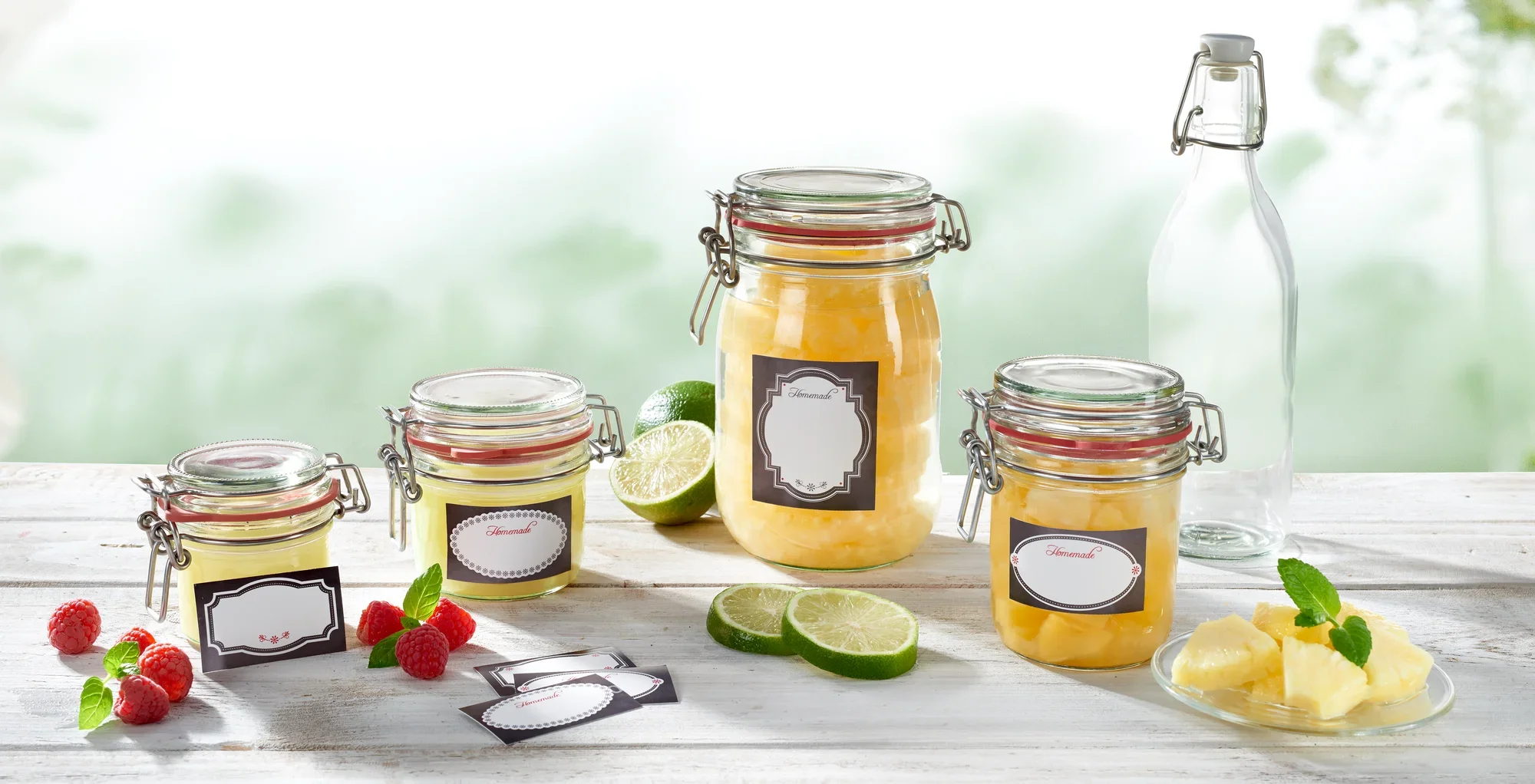Vier inmaakpotten met decoratieve zwart-witte etiketten staan op een tafel. Ze zijn gevuld met gele inhoud, omringd door limoenen, frambozen en stukjes ananas.