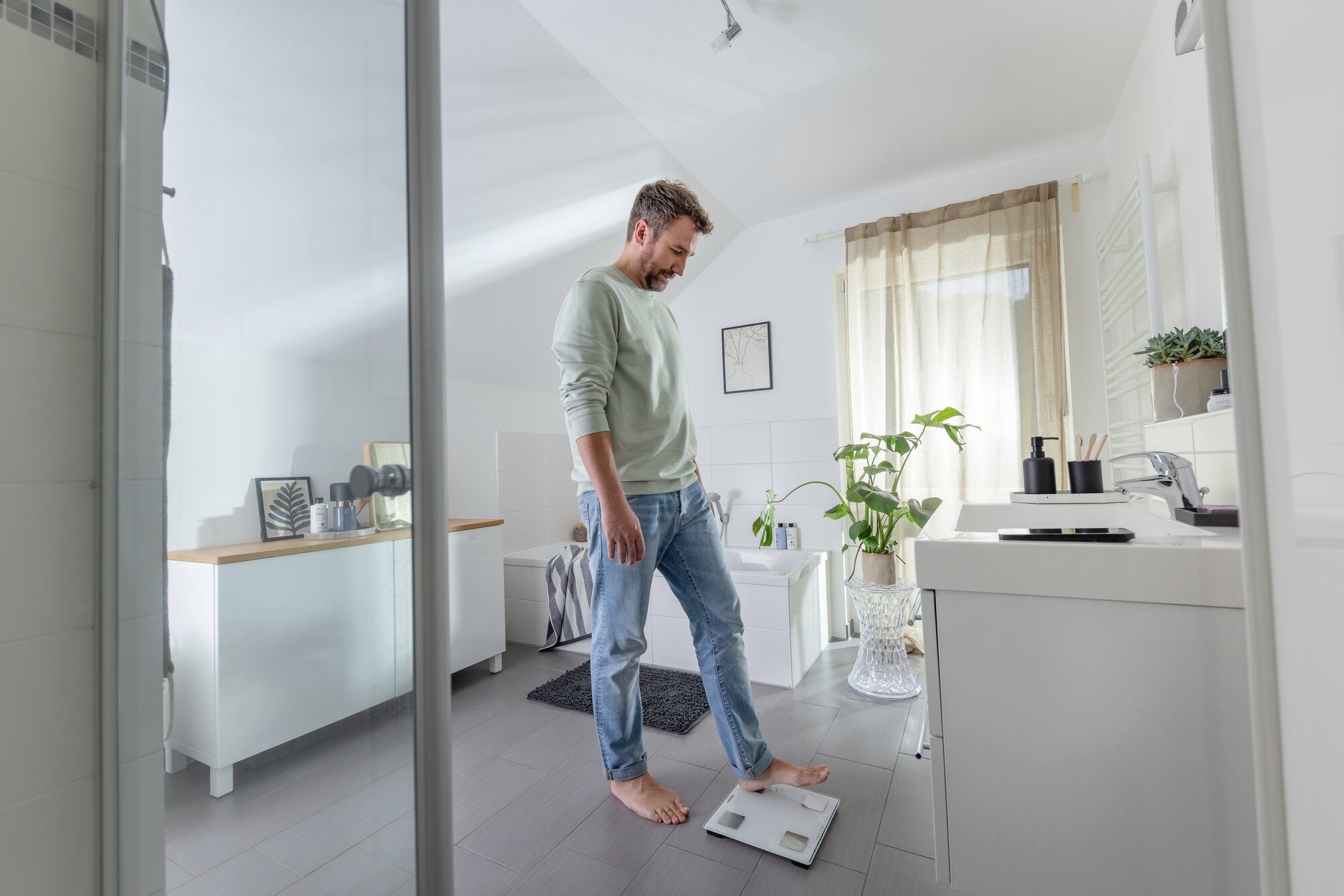A man is standing in a modern bathroom on a digital bathroom scale, model Shape Sense Connect Lite. The surroundings are bright and minimalist.