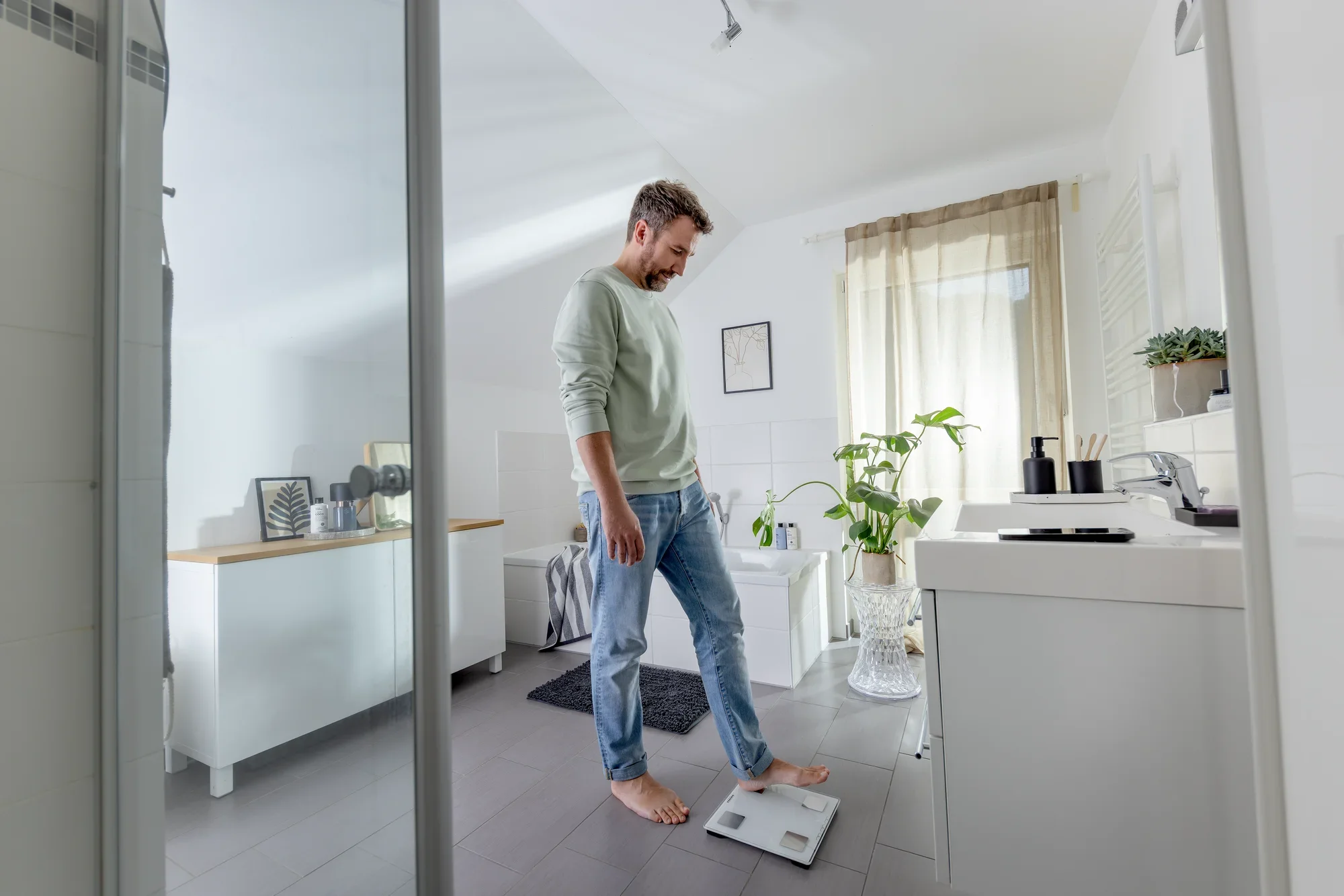 Mężczyzna stoi na cyfrowej wadze łazienkowej, modelu Shape Sense Connect Lite, w nowoczesnej łazience. Otoczenie jest jasne i minimalistyczne.