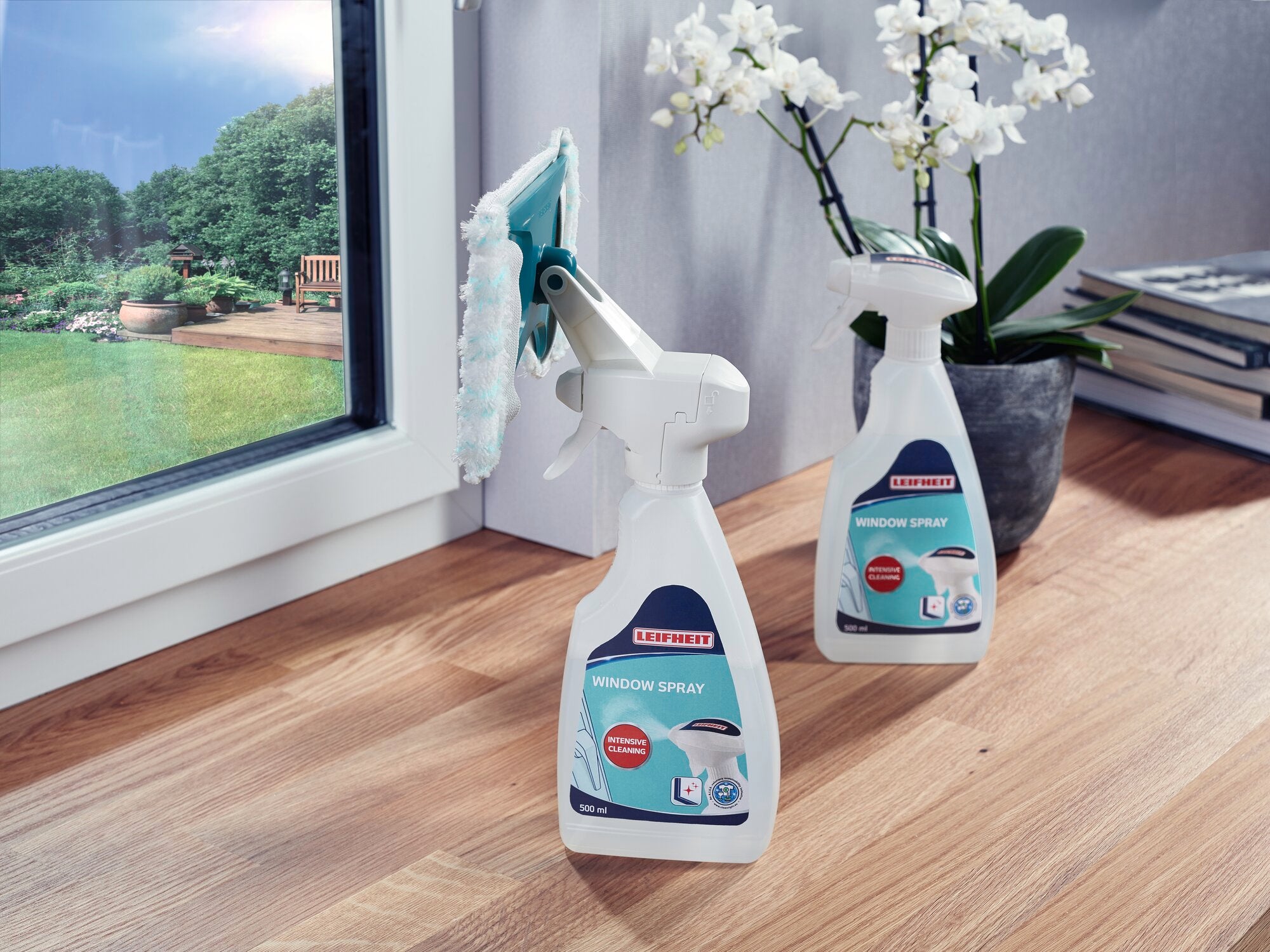 Zwei Flaschen des Leifheit Window Spray Cleaners stehen auf einem Holzboden neben einem Fenster. Eine Flasche ist mit einem Sprühwischer ausgestattet. Im Hintergrund sind Pflanzen sichtbar.