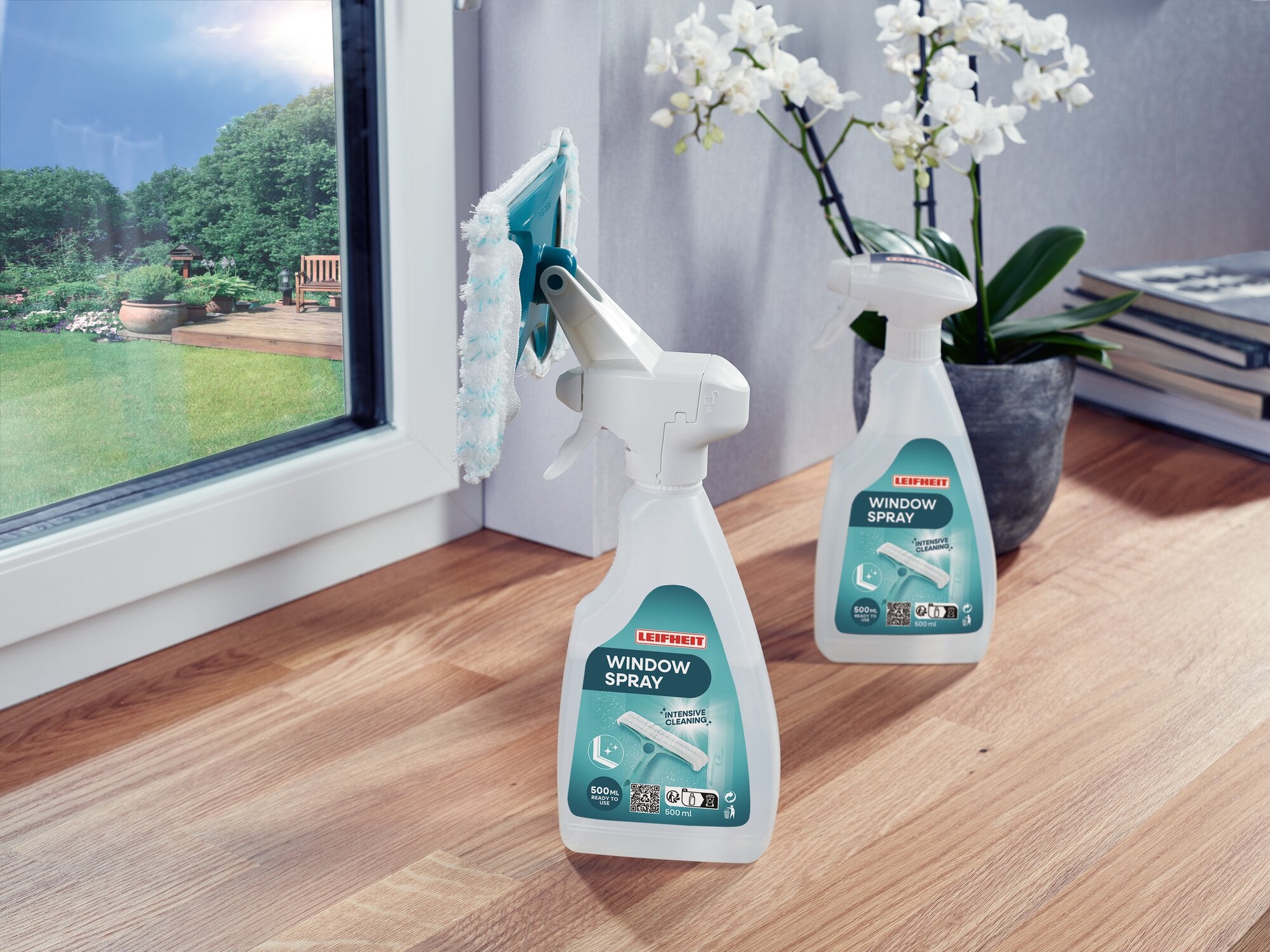 Zwei Flaschen des Leifheit Window Spray Cleaners stehen auf einem Holzboden neben einem Fenster. Eine Flasche ist mit einem Sprühwischer ausgestattet. Im Hintergrund sind Pflanzen sichtbar.