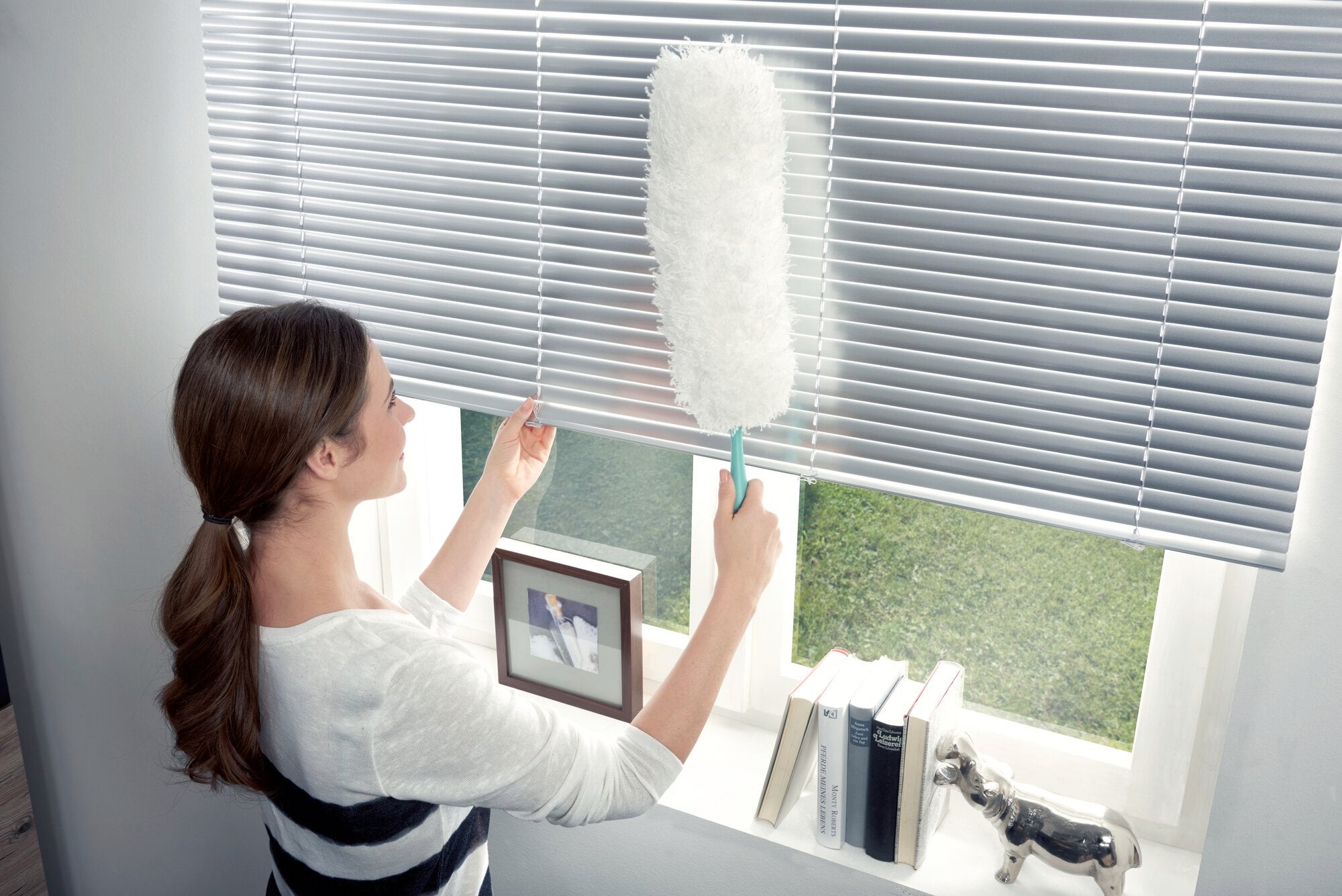 A woman uses the feather duster to remove dust from blinds. The feather duster is white and fluffy, ideal for hard-to-reach places. Books and a window can be seen in the background.