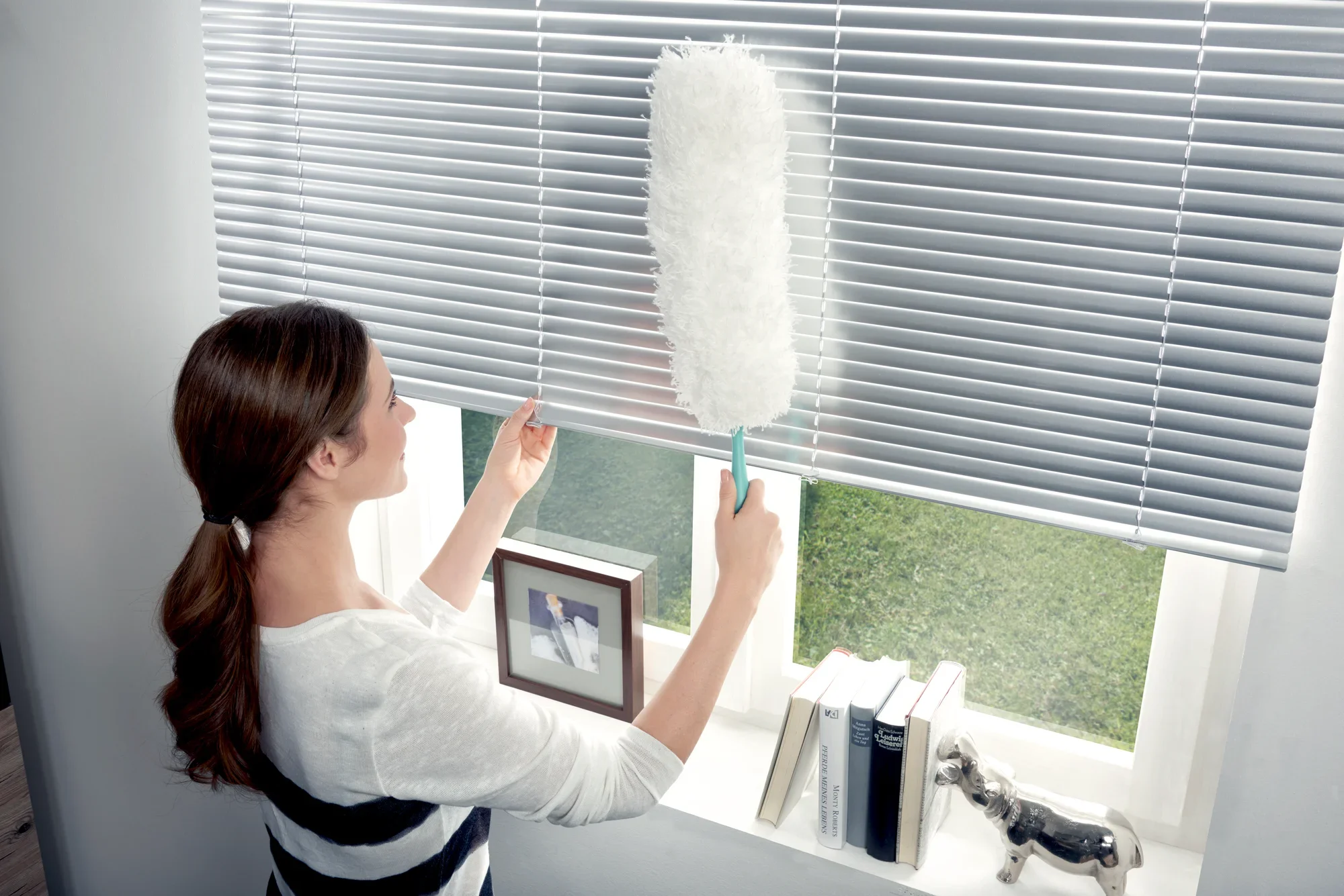Una mujer utiliza el plumero para quitar el polvo de las persianas. El plumero es blanco y mullido, ideal para lugares de difícil acceso. Al fondo se ven libros y una ventana.