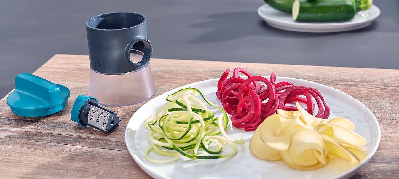 Ein Spiralschneider mit Klinge und Deckel, daneben ein Teller mit spiralfoermigen Gemuesen aus Zucchini, roter Bete und Kartoffeln.