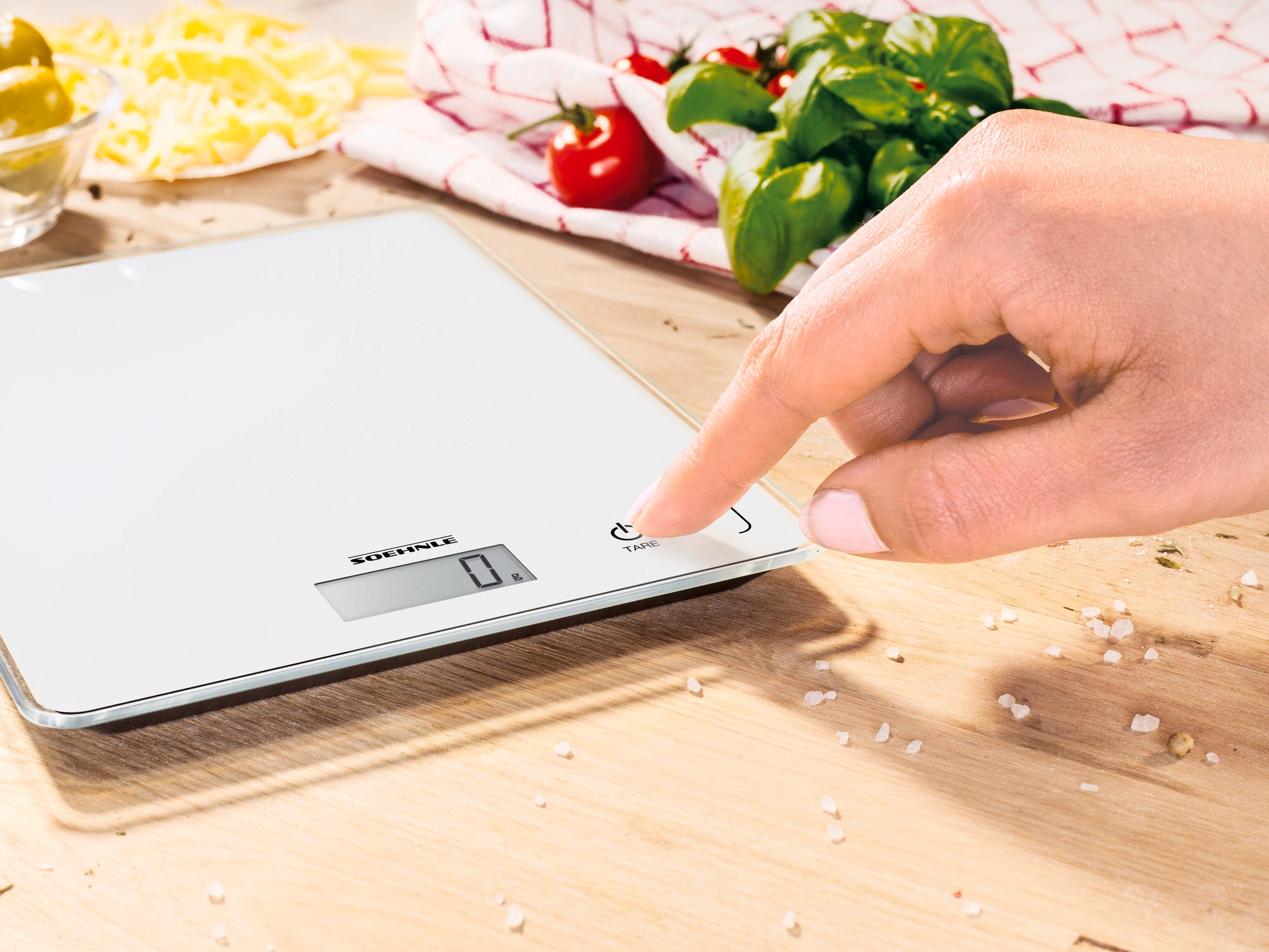 Een digitale keukenweegschaal, model Page Compact 300, wordt gebruikt op een houten werkblad. Een hand drukt op de inschakelknop. Op de achtergrond zijn verse ingrediënten te zien.