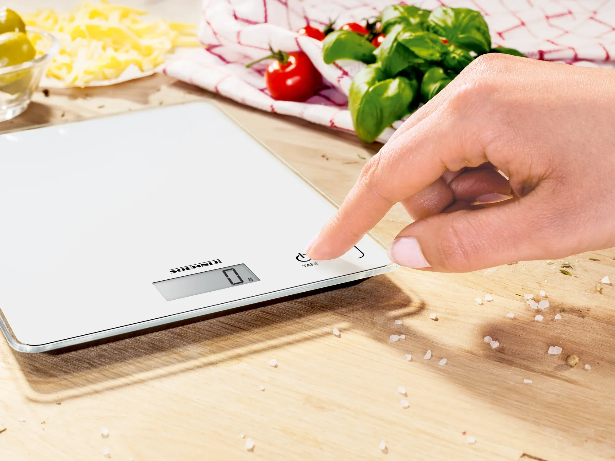 A digital kitchen scale, model Page Compact 300, is being used on a wooden surface. A hand presses the power button. Fresh ingredients can be seen in the background.