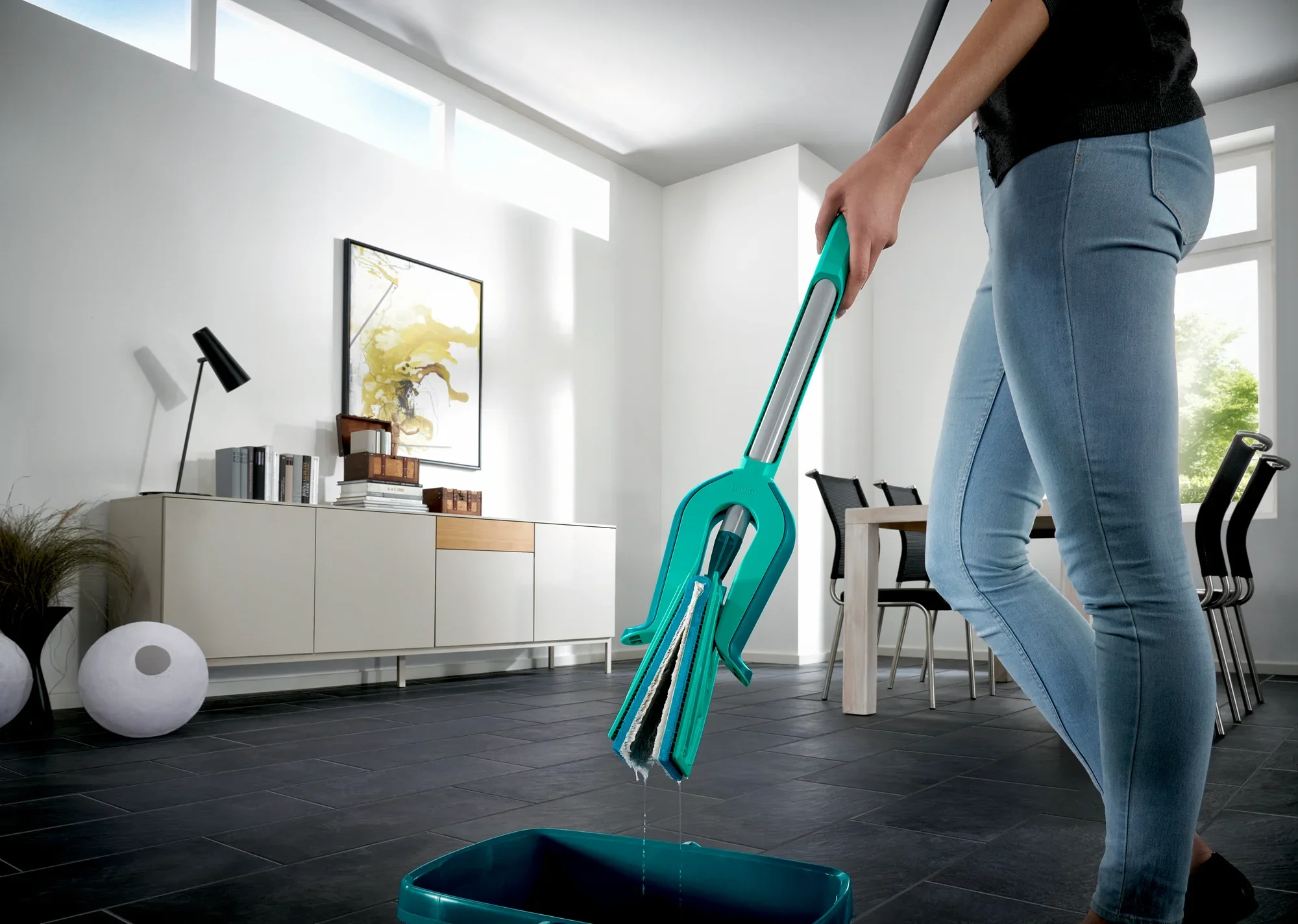 Una persona limpia el suelo con el fregona Picobello XL micro duo en un salón moderno. La fregona tiene un mango ergonómico y un sistema de limpieza eficaz. Ideal para una limpieza a fondo de baldosas y laminados.