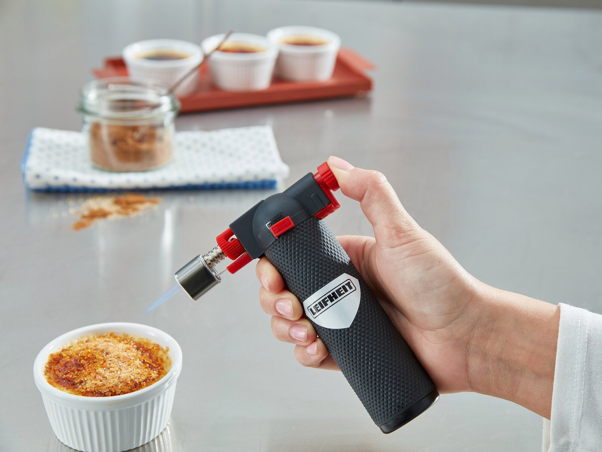 Eine Hand hält einen Flambierer von Leifheit, während eine Crème brûlée in einer weißen Schale flambiert wird. Im Hintergrund sind weitere Schalen und ein Glas mit Zucker zu sehen.