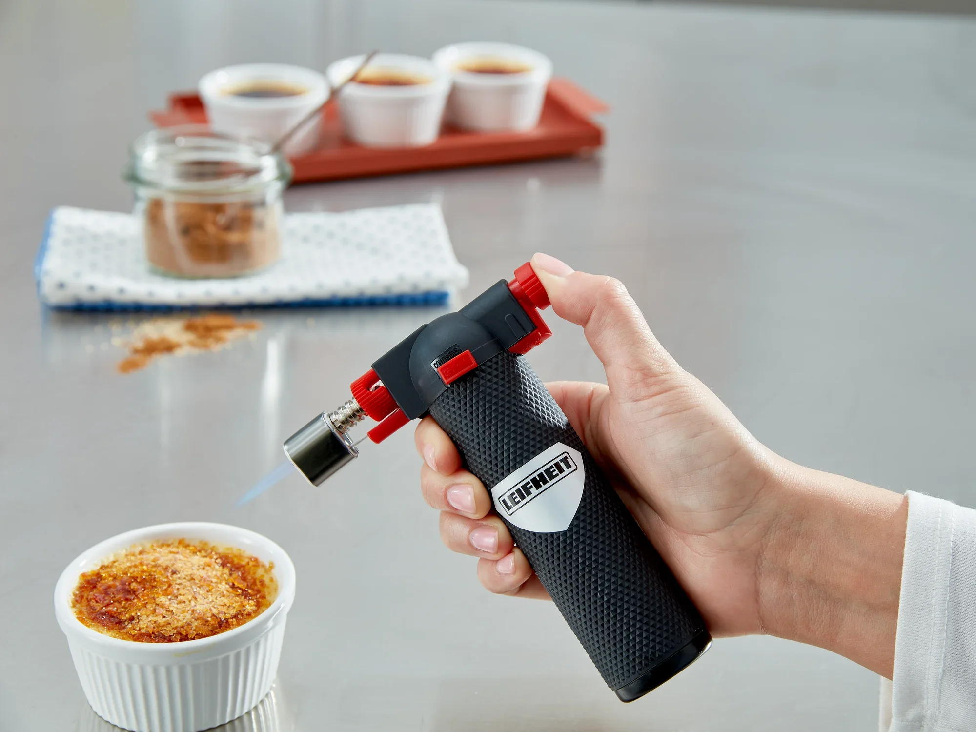 Eine Hand hält einen Flambierer von Leifheit, während eine Crème brûlée in einer weißen Schale flambiert wird. Im Hintergrund sind weitere Schalen und ein Glas mit Zucker zu sehen.