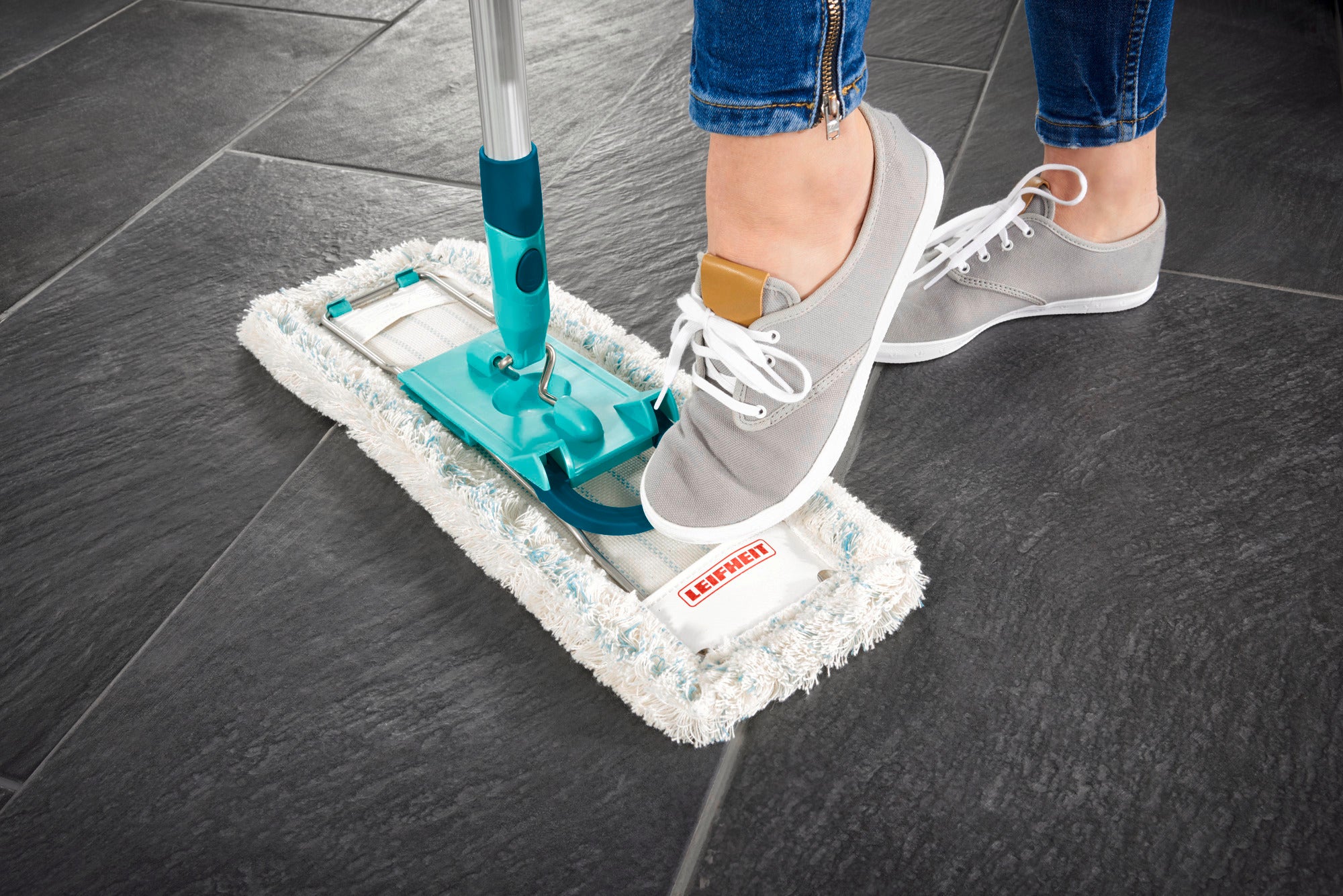 Un balai Profi XL est utilisé sur un carrelage foncé. Une personne chaussée de chaussures grises actionne le mécanisme de balayage avec son pied pour nettoyer le balai.