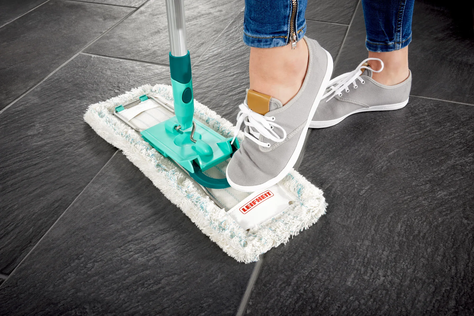 Un lavapavimenti Profi XL viene utilizzato su piastrelle scure. Una persona con scarpe grigie aziona il meccanismo di lavaggio con il piede per pulire il lavapavimenti.