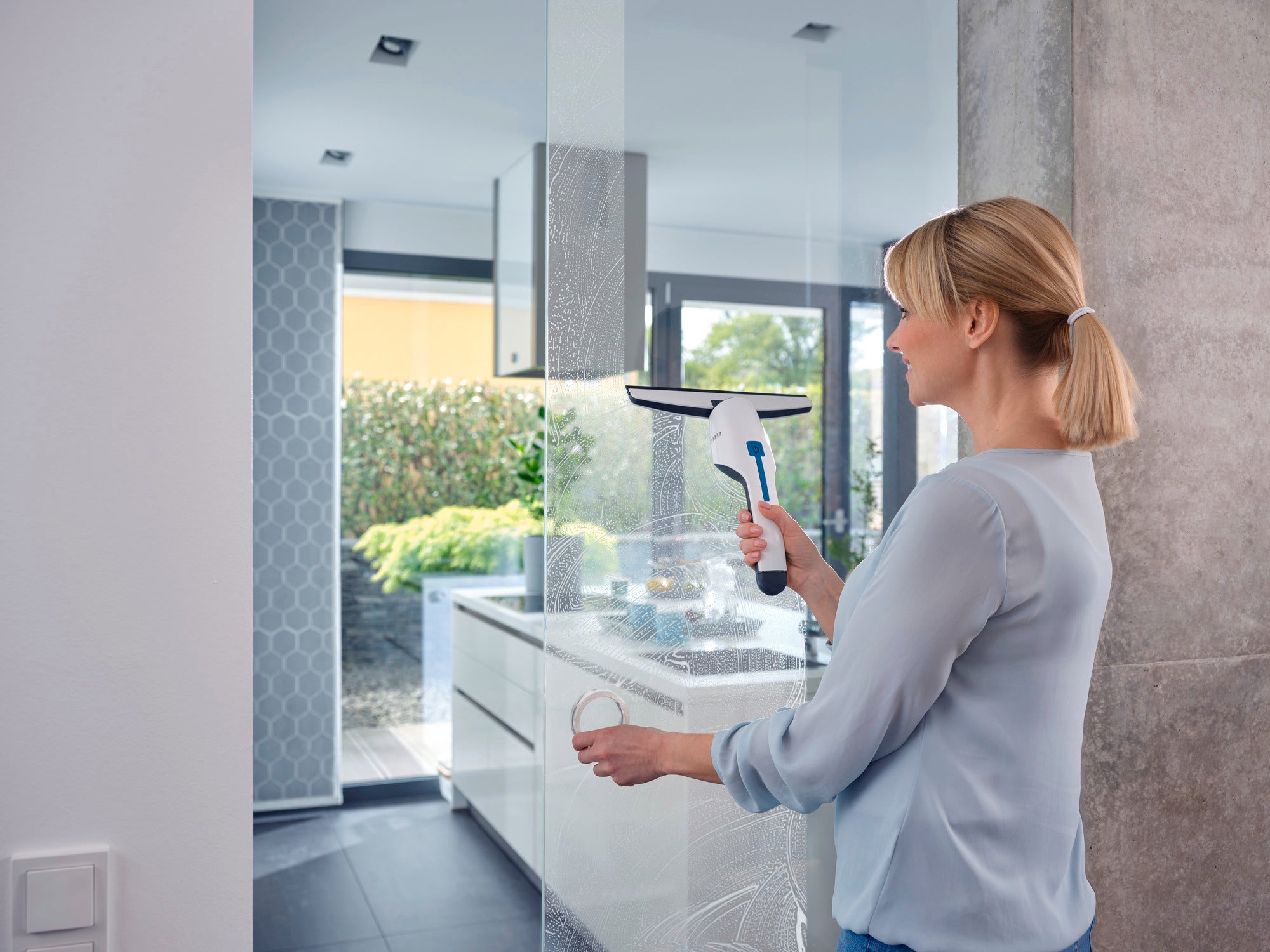 Una mujer limpia una ventana grande en una cocina moderna con el aspirador para ventanas Nemo. El aspirador es compacto y eficiente, ideal para una limpieza sin rayas.