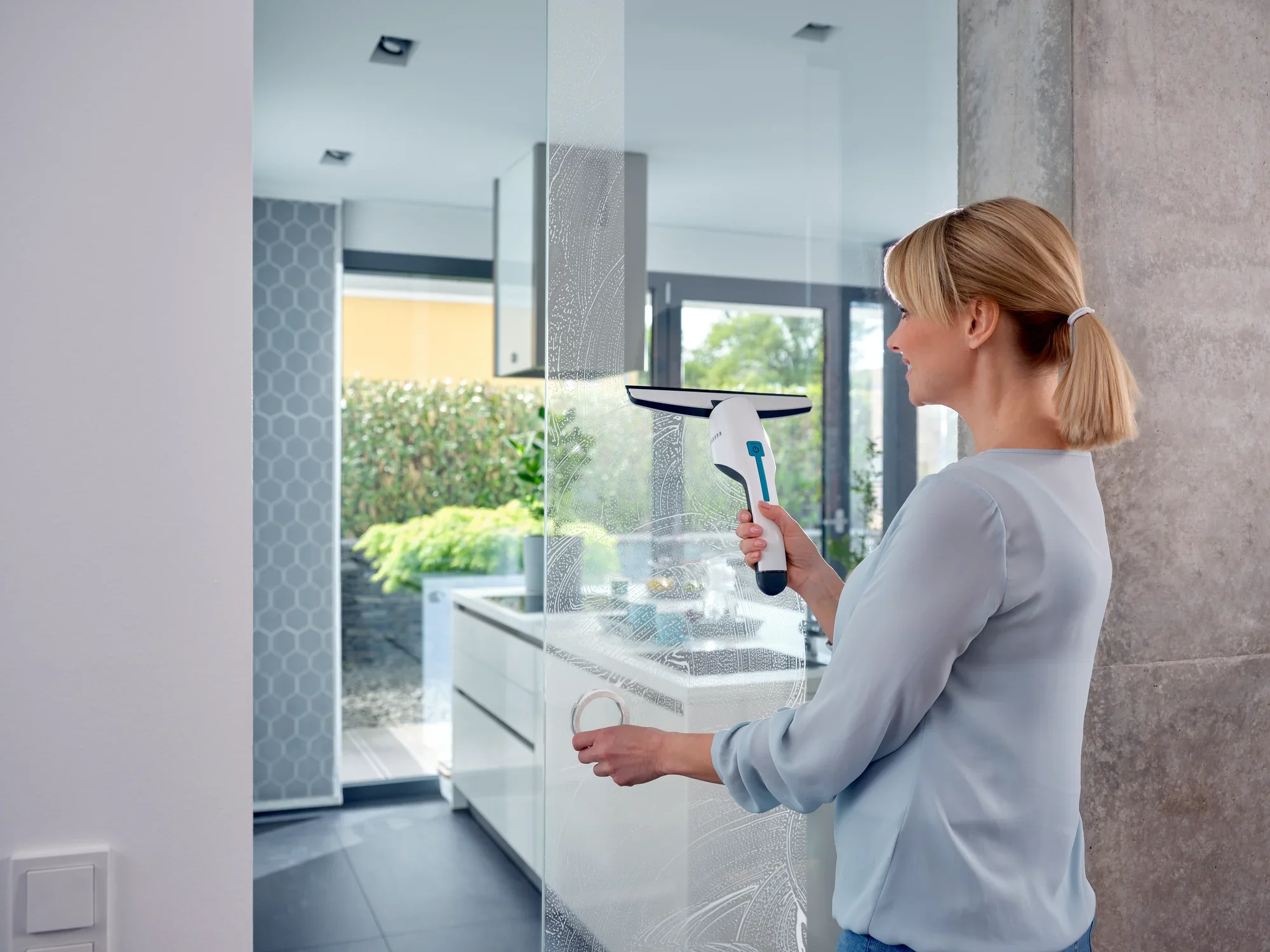 Une femme nettoie une grande fenêtre dans une cuisine moderne avec le nettoyeur de vitres Nemo. Le nettoyeur est compact et efficace, idéal pour une propreté sans traces.