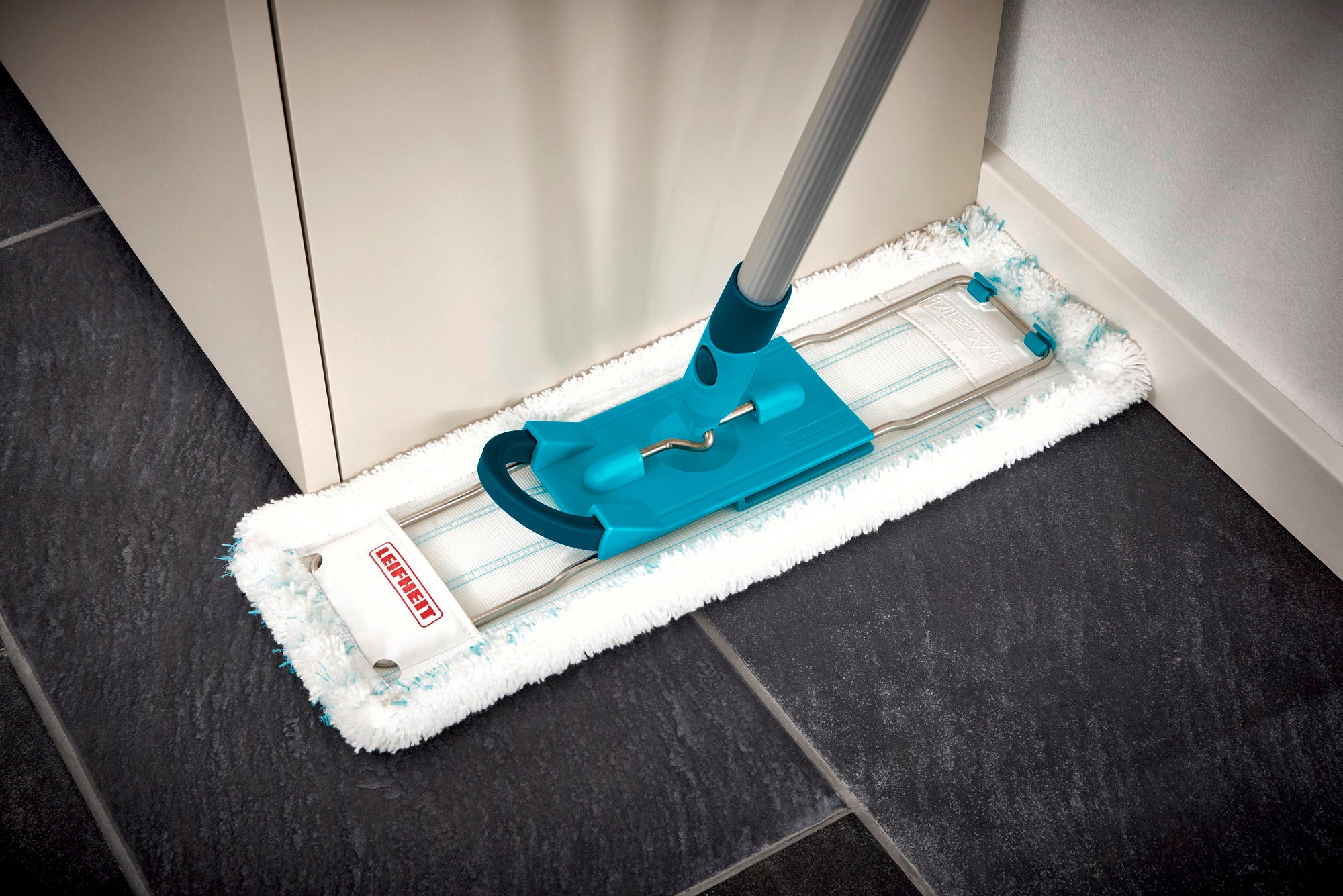 Ein Bodenwischer Profi XL mit einem blauen Griff reinigt eine dunkle Fliesenfläche in der Ecke eines Raumes. Der Wischbezug ist weiß und flauschig, ideal für gründliche Reinigung.