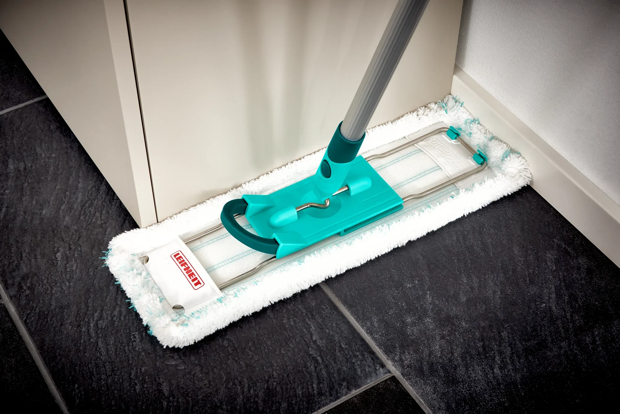 A Profi XL floor mop with a blue handle cleans a dark tiled surface in the corner of a room. The mop cover is white and fluffy, ideal for thorough cleaning.