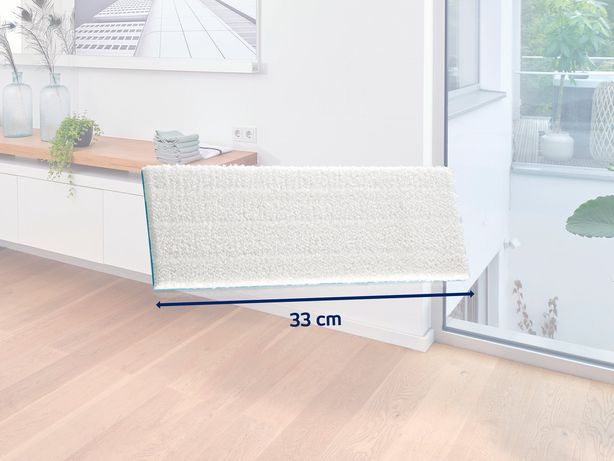Une serpillière blanche d'une largeur de 33 cm repose sur un sol en bois clair dans une pièce moderne. À l'arrière-plan, on aperçoit un buffet décoré et une grande fenêtre.