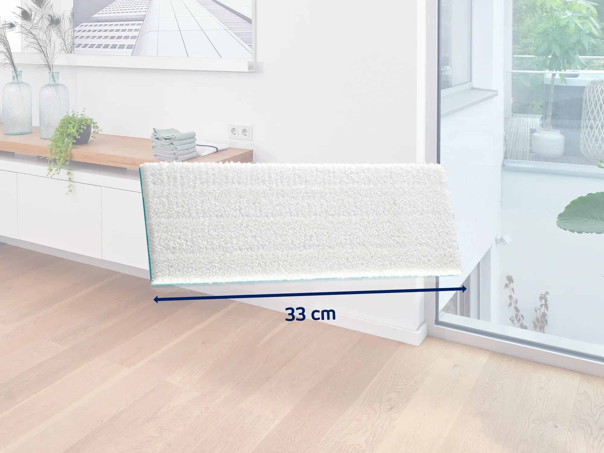 A white mop with a width of 33 cm lies on a light wooden floor in a modern room. In the background, you can see a sideboard with decorations and a large window.