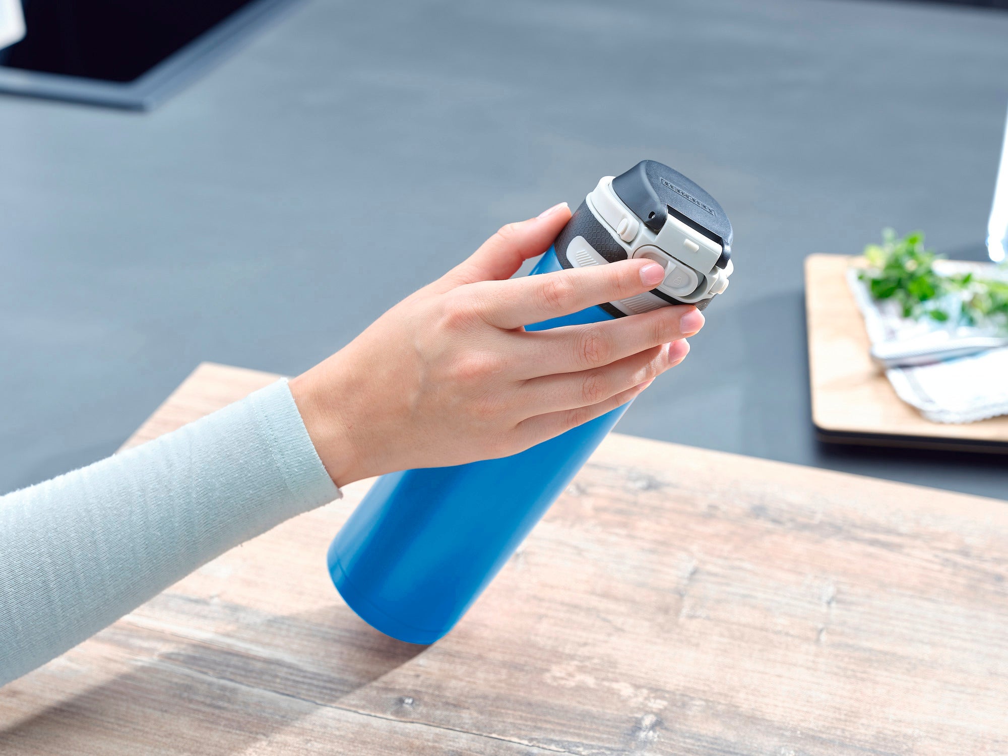 Ein Isolierbecher Flip in Hellblau mit 600 ml Fassungsvermögen wird auf einem Tisch gehalten. Der Becher hat einen schwarzen Deckel und liegt in einer Hand. Im Hintergrund sind Küchenutensilien zu sehen.