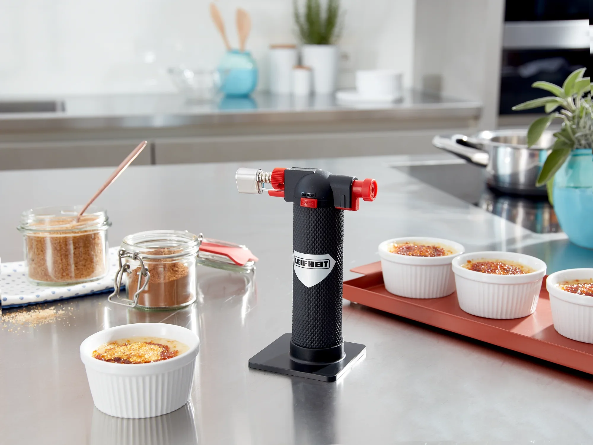 Ein Flambierer von Leifheit steht auf einer Küchenarbeitsplatte, umgeben von Crème Brûlée in weißen Schälchen. Im Hintergrund sind Küchenutensilien und ein Topf zu sehen.