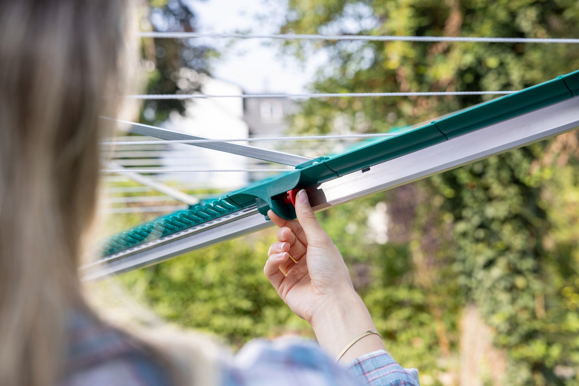 Eine Person bedient die Leifheit Wäschespinne Linomatic 600 Deluxe im Garten. Die Hand zieht an einem grünen Hebel, um die Leinen zu spannen. Im Hintergrund sind Bäume und Büsche zu sehen.