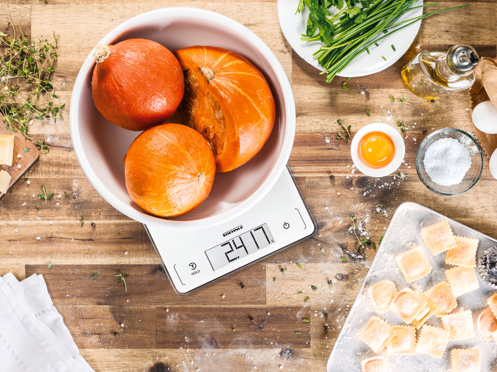 La balance de cuisine numérique Page Profi 300 affiche 2471 grammes. Un saladier contenant des courges est posé sur la balance, entouré d'herbes fraîches, d'huile et de raviolis sur une planche en bois.