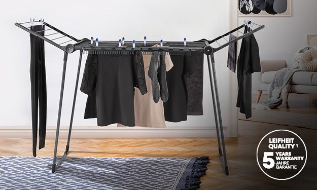 Ein schwarzgrauer Waeschestaender steht in einem modern eingerichteten Wohnzimmer mit Parkettboden, behaengt mit schwarzer und beiger Kleidung.