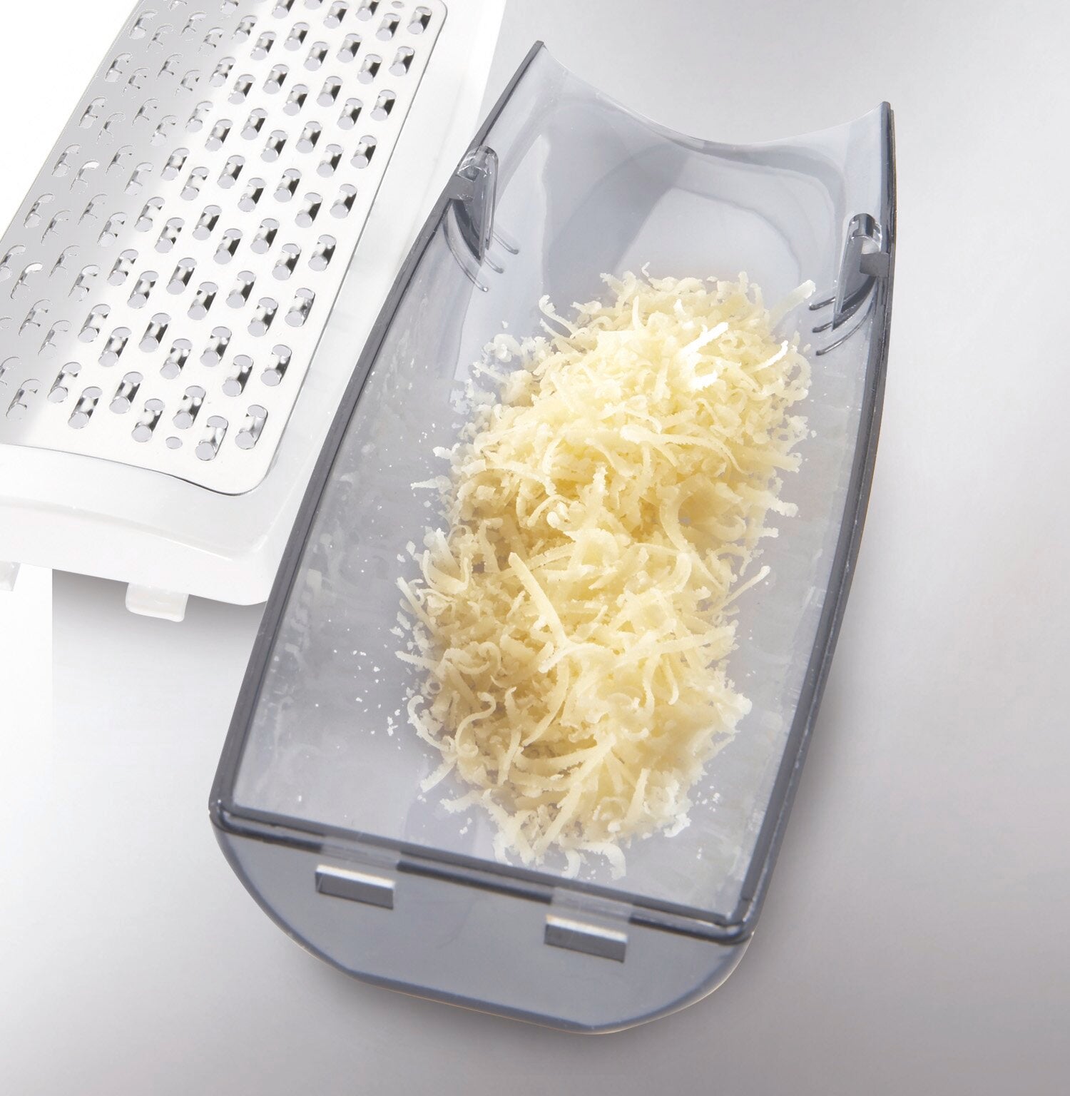 Râpe à parmesan en action : un bol transparent contenant du parmesan fraîchement râpé. À l'arrière-plan, on aperçoit la râpe métallique qui a râpé le fromage.