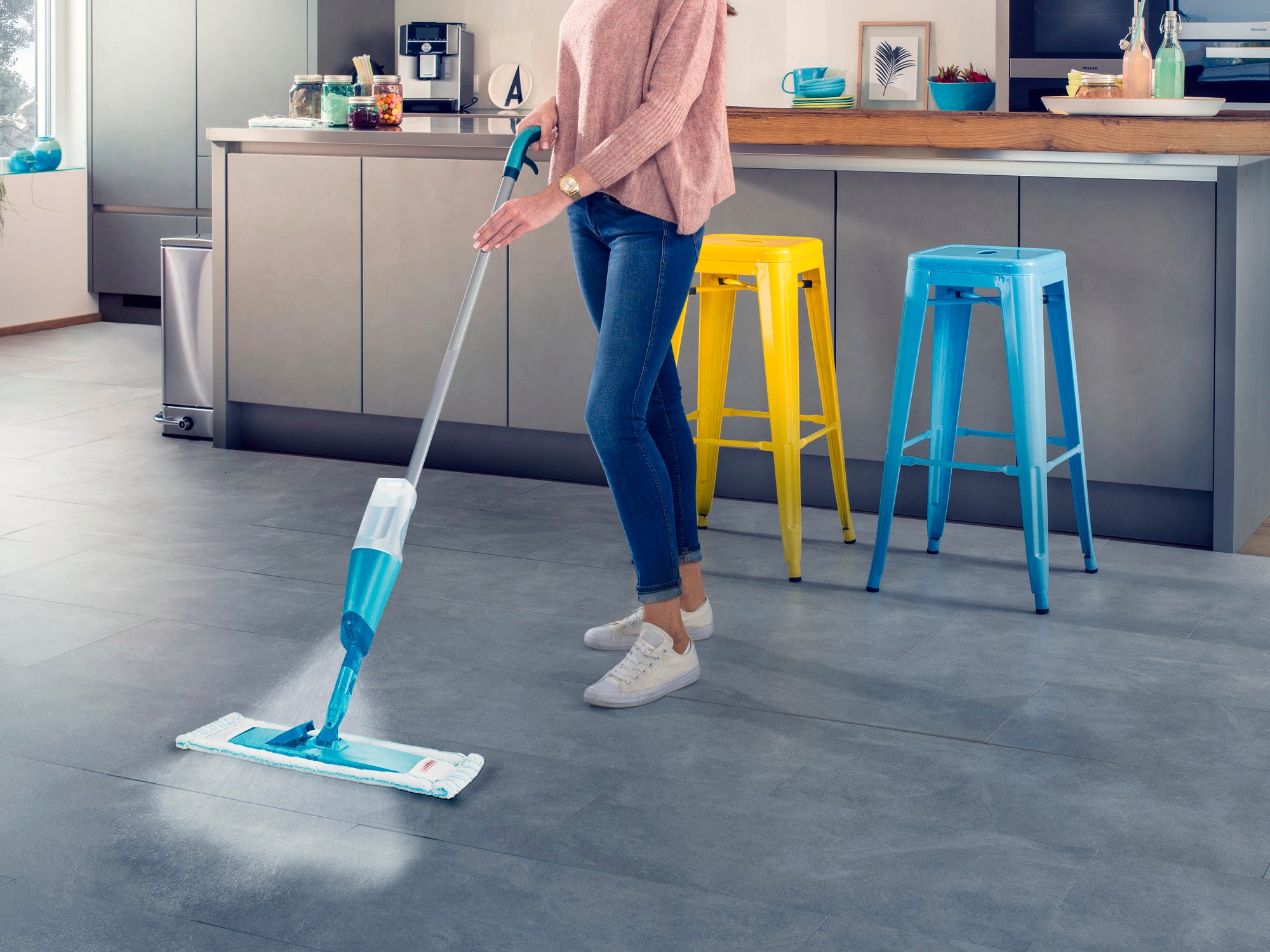 Une personne nettoie le sol de la cuisine avec le balai à franges Easy Spray XL. Le balai pulvérise de l'eau sur le sol tandis que la personne le passe sans effort sur le carrelage. Des tabourets de bar colorés sont visibles à l'arrière-plan.