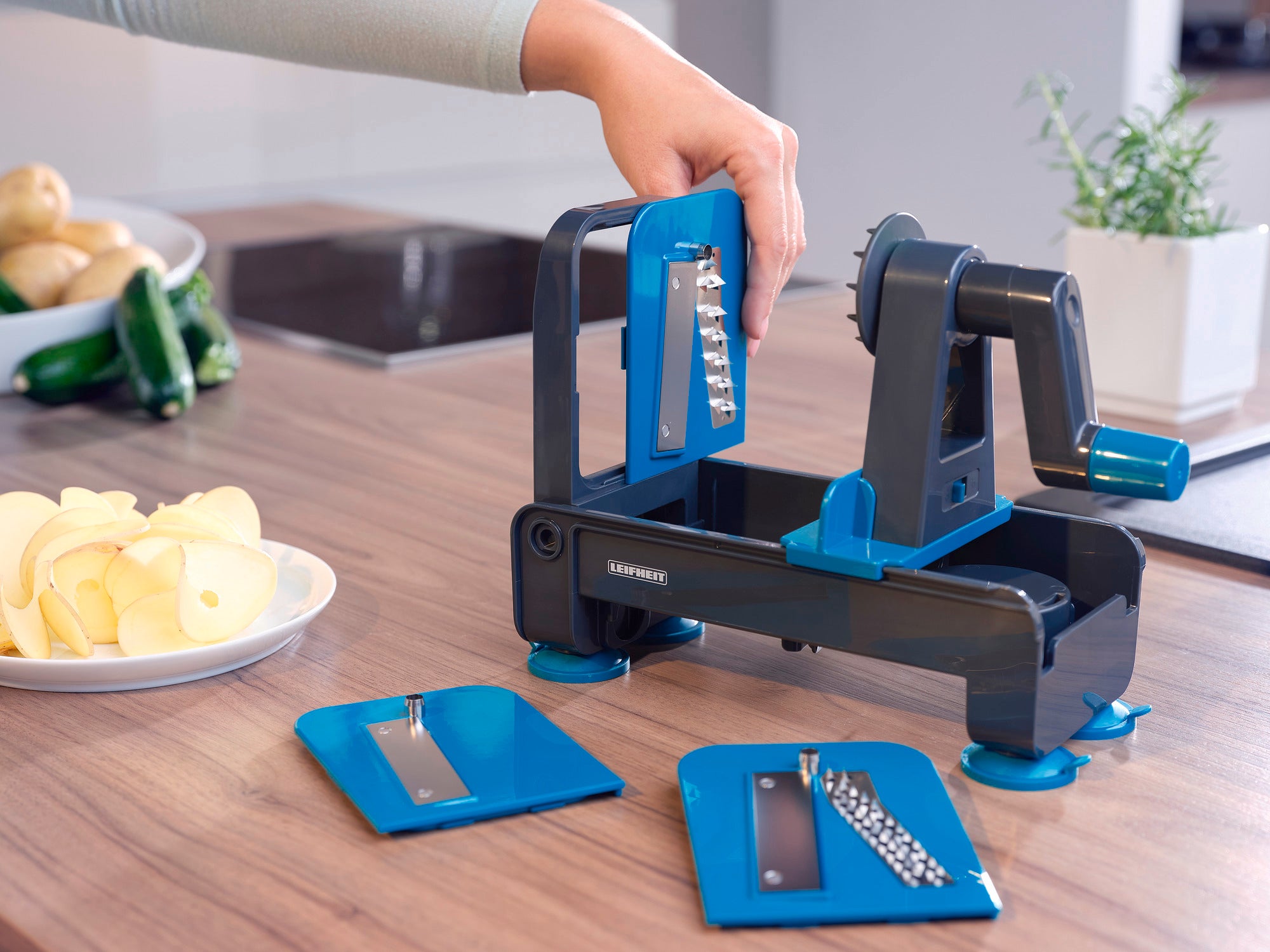 Een tafelspiraalsnijder wordt gebruikt op een keukenblad. Eén hand bedient het apparaat terwijl op de achtergrond gesneden appels en courgettes te zien zijn.