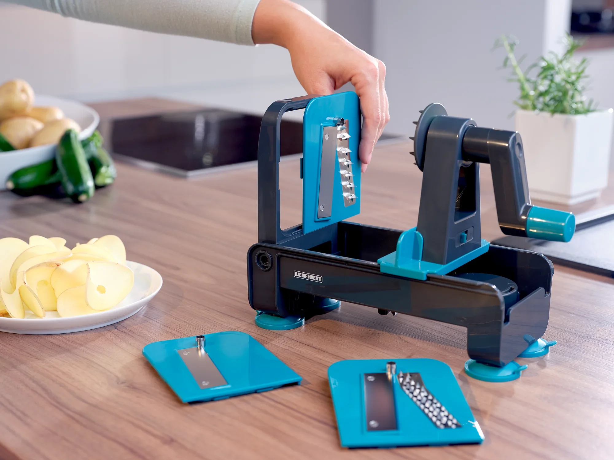 Ein Tisch-Spiralschneider wird auf einer Küchenarbeitsplatte verwendet. Eine Hand bedient das Gerät, während geschnittene Äpfel und Zucchini im Hintergrund zu sehen sind.