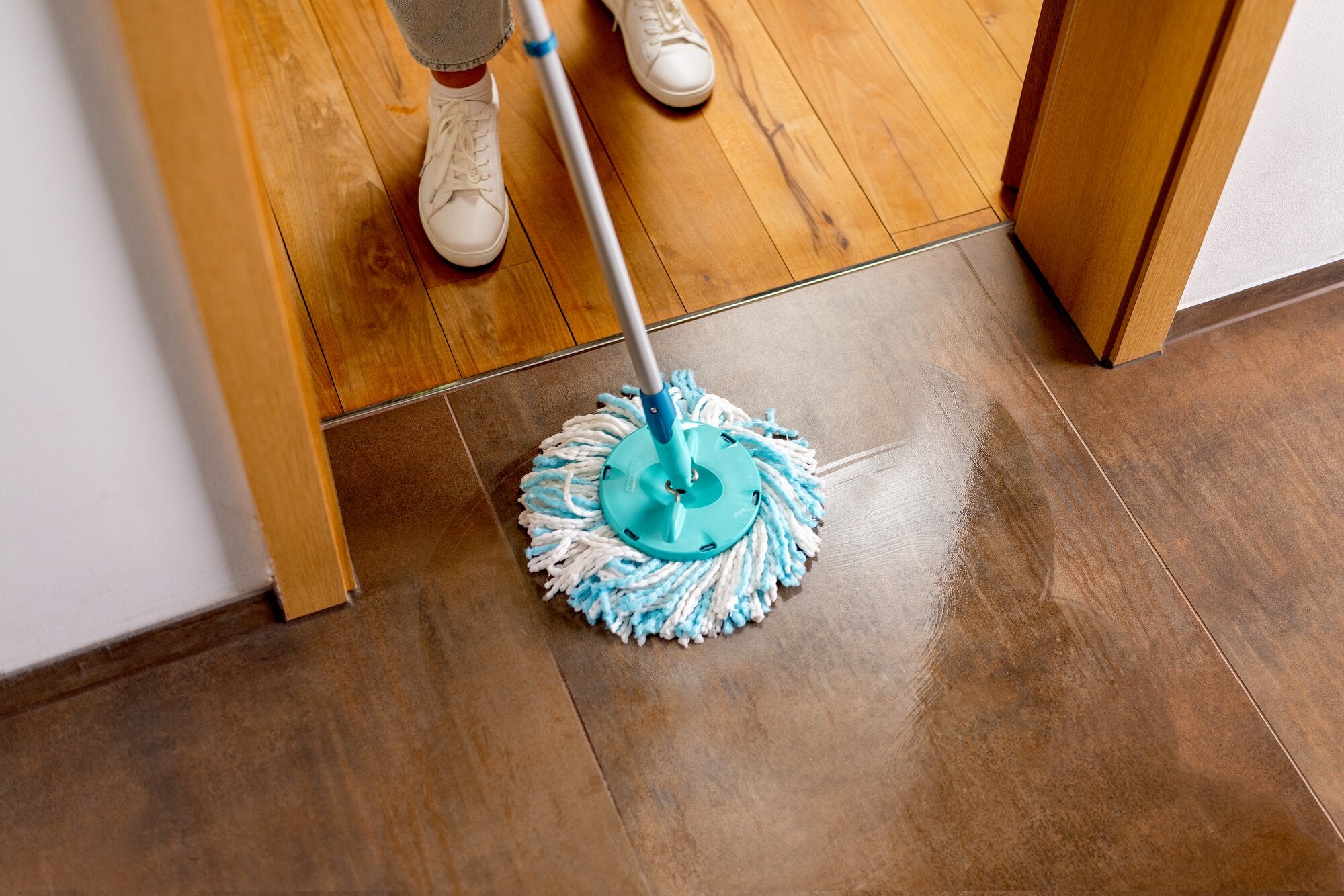 Leifheit CLEAN TWIST Disc Mop reinigt een betegelde vloer in een deuropening, waarbij de turquoise ronde mopkop hoeken en randen bereikt en vuil verwijdert van bruine tegels naast een houten vloer.