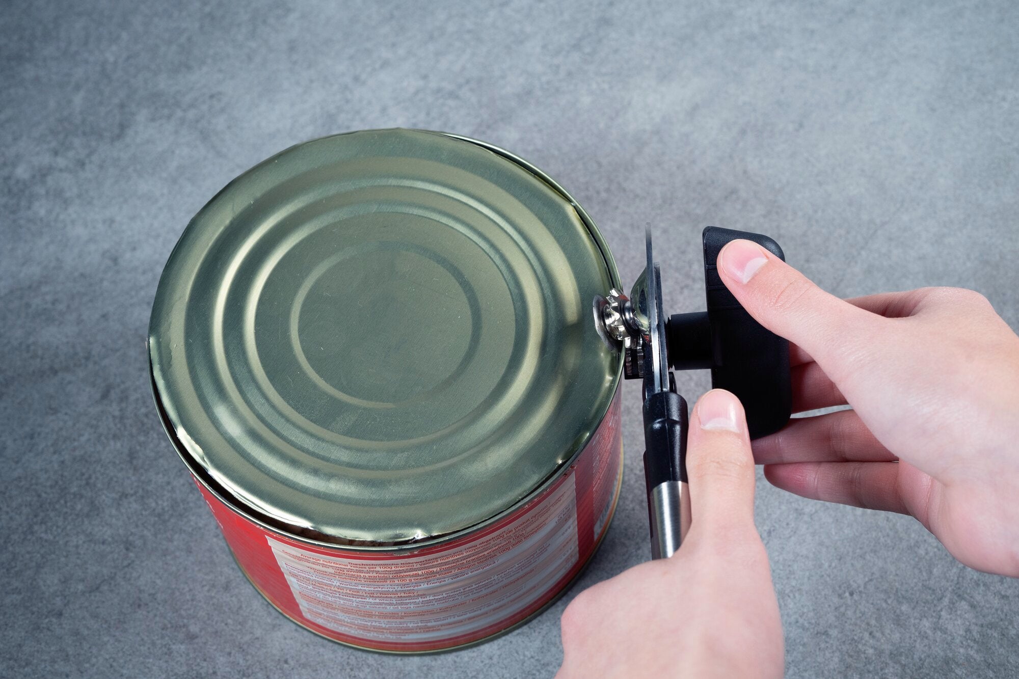 Een hand opent een groot, rond blik met een zwart handvat blikopener op een grijze ondergrond.