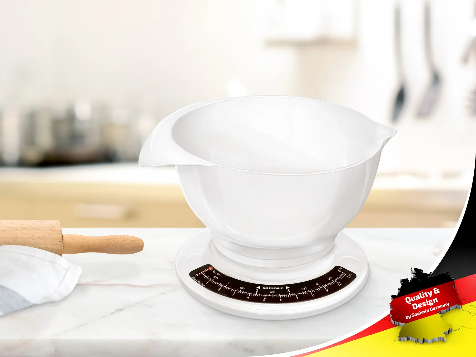 Biała waga kuchenna z wyjmowaną miską stoi na blacie kuchennym. W tle widoczne są przybory kuchenne. Znak jakości znajduje się w prawym dolnym rogu zdjęcia.