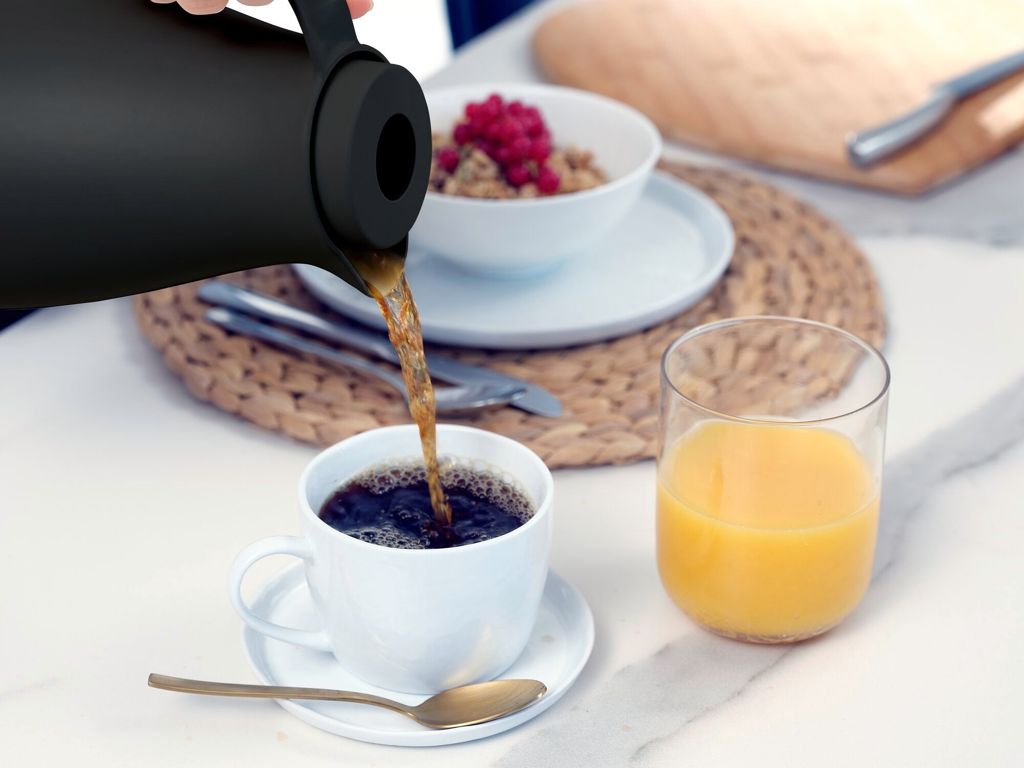 Eine schwarze Isolierkanne namens Harmonic gießt heißen Kaffee in eine weiße Tasse. Daneben stehen ein Glas Orangensaft und eine Schale mit Müsli auf einem Tisch.