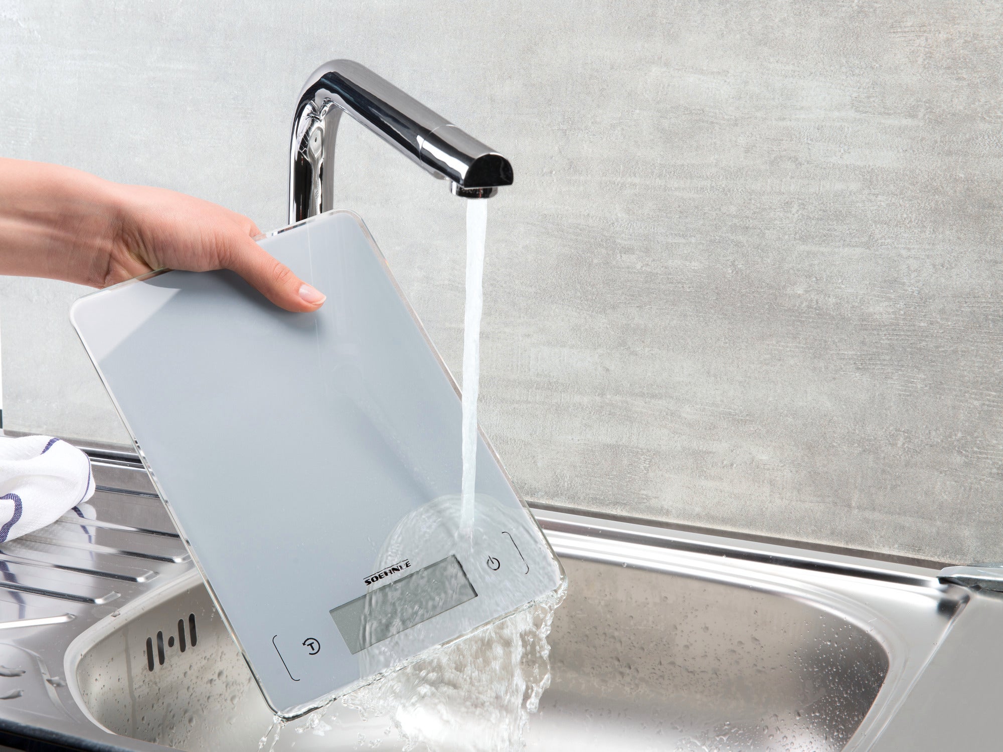 Digitale Küchenwaage Page Aqua Proof wird unter fließendem Wasser gereinigt. Die Waage ist wasserdicht und ideal für den Einsatz in der Küche geeignet.