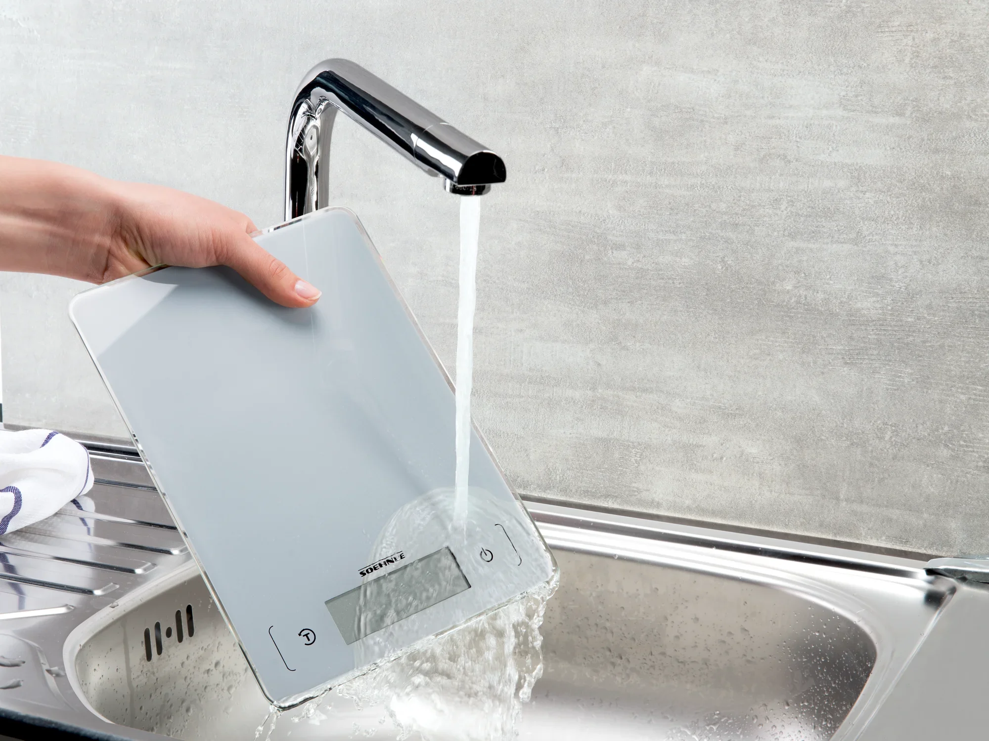 Digitale Küchenwaage Page Aqua Proof wird unter fließendem Wasser gereinigt. Die Waage ist wasserdicht und ideal für den Einsatz in der Küche geeignet.