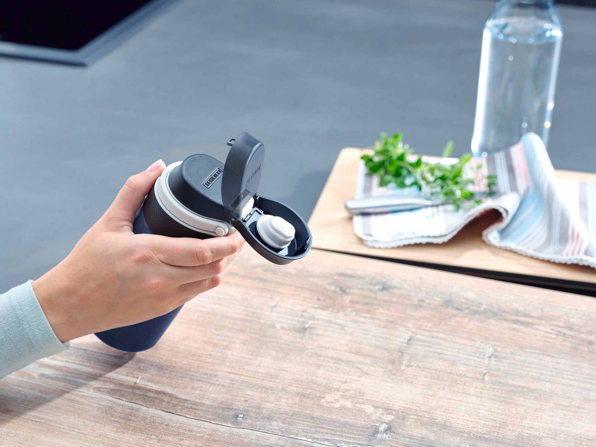 Una persona tiene in mano un thermos Flip blu scuro da 350 ml. Il thermos ha il coperchio aperto. Sullo sfondo si vedono un panno, delle erbe aromatiche e una bottiglia d'acqua.