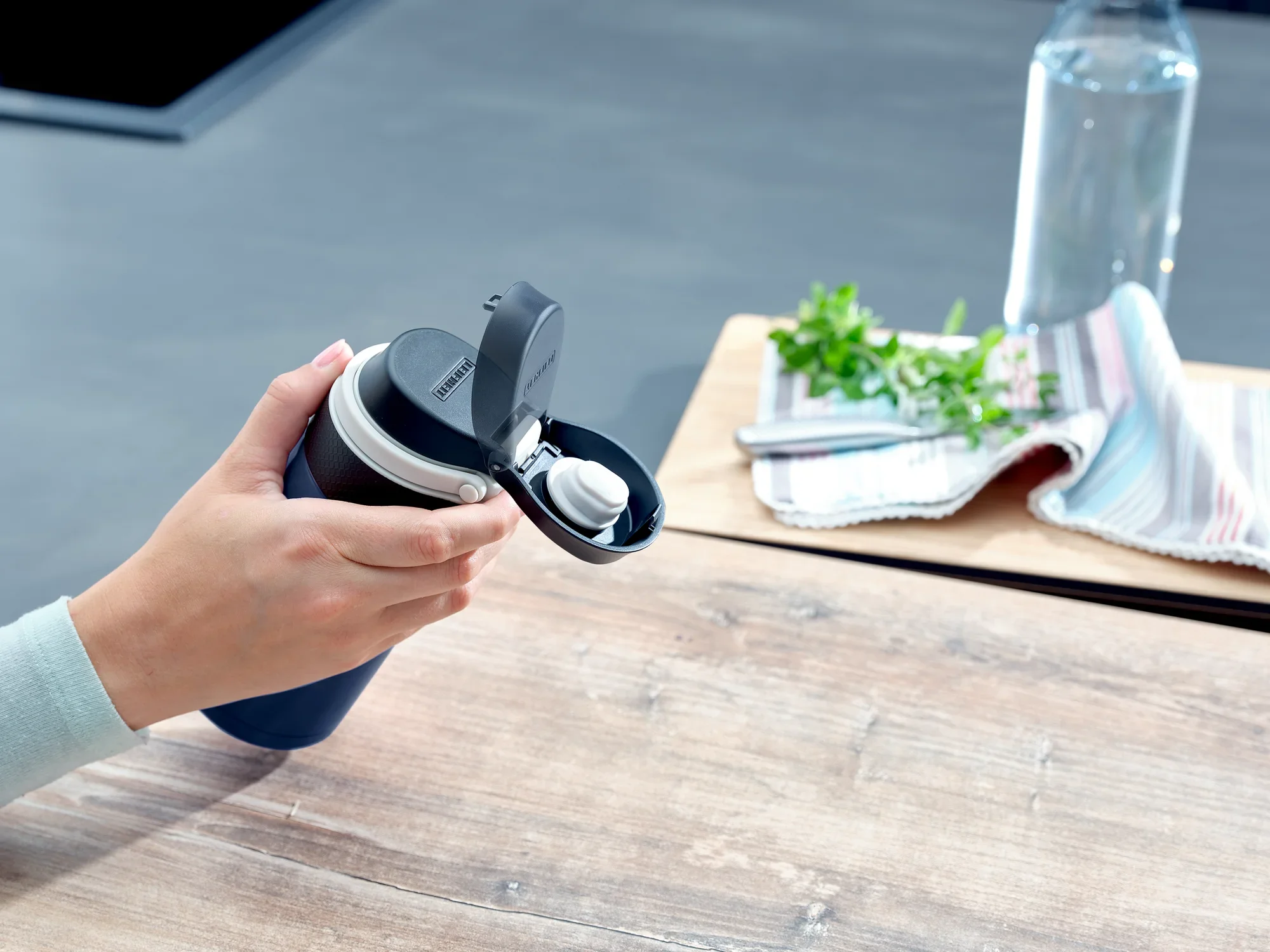 Osoba trzyma ciemnoniebieski kubek próżniowy Flip o pojemności 350 ml. Kubek ma otwartą pokrywkę. W tle widać ściereczkę, zioła i butelkę z wodą.