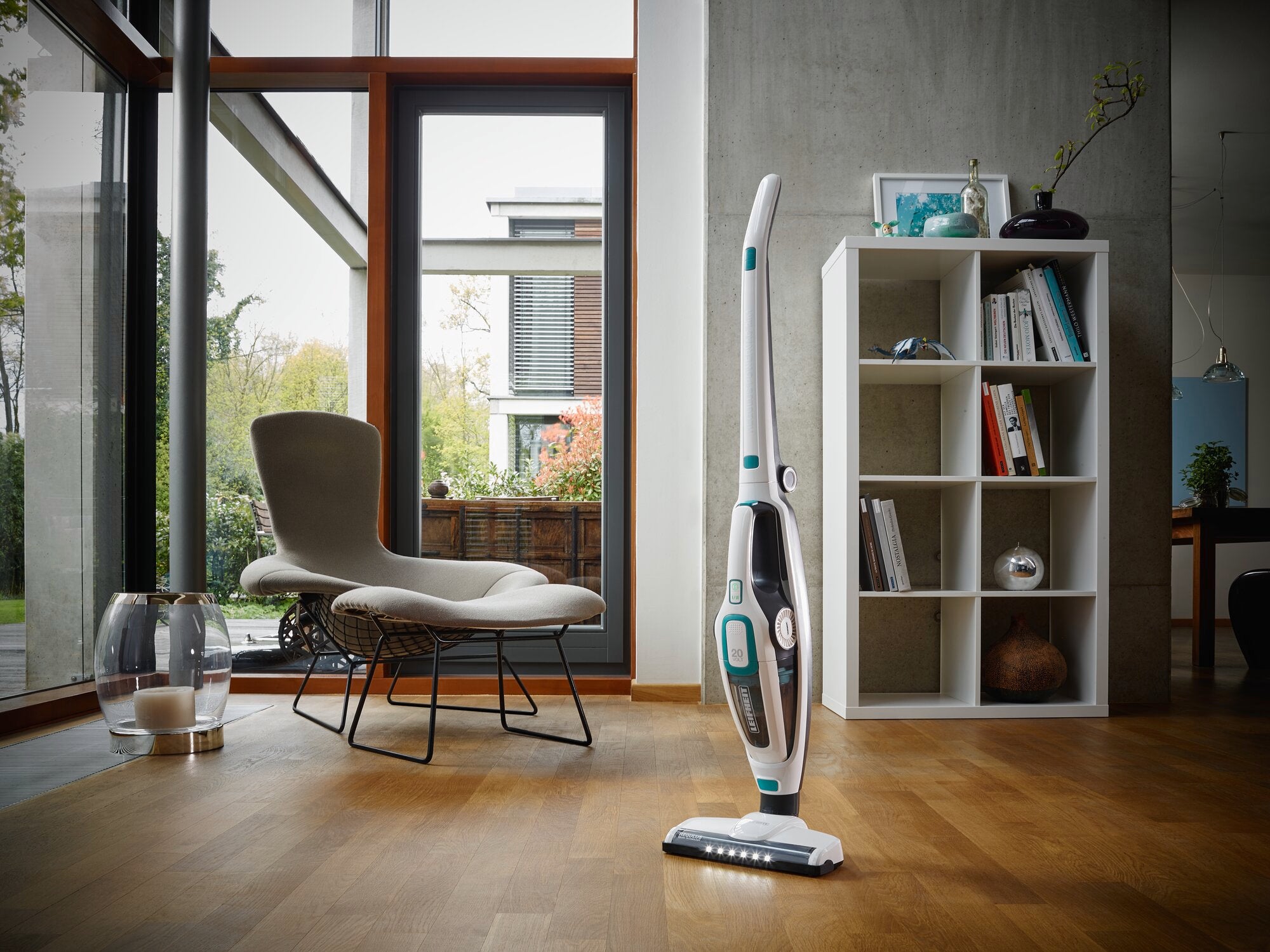 Ein stilvoller, moderner Raum mit einem Regulus PowerVac 2in1 Staubsauger auf einem Holzboden. Im Hintergrund sind ein Sessel, ein Regal und große Fenster zu sehen.