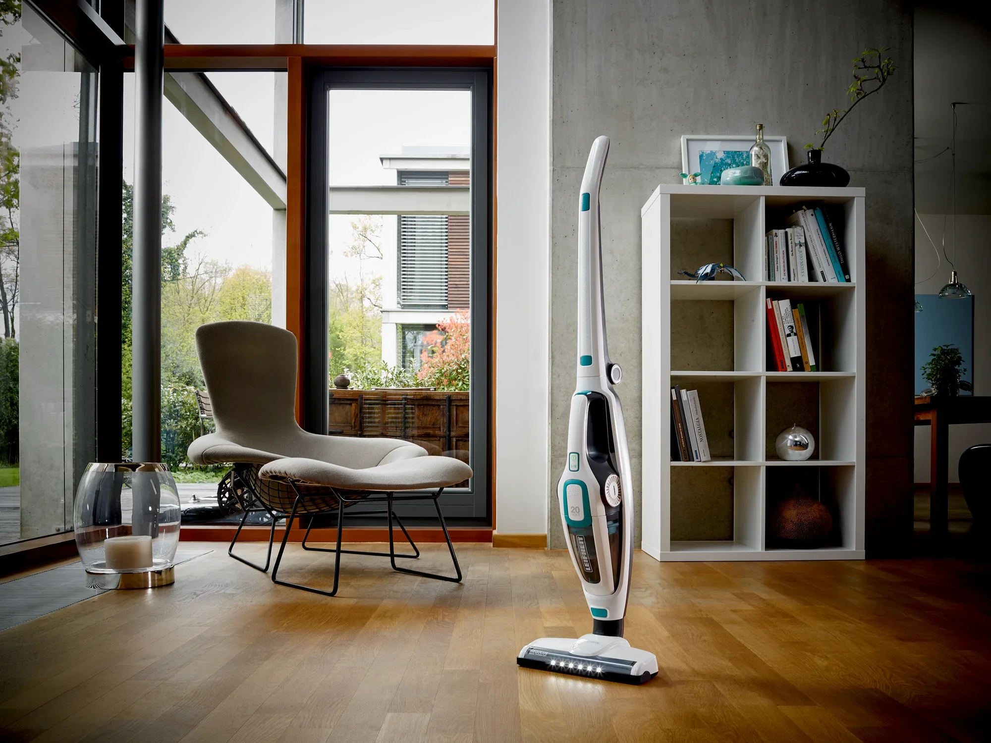 Une pièce élégante et moderne avec un aspirateur Regulus PowerVac 2 en 1 sur un sol en bois. À l'arrière-plan, on aperçoit un fauteuil, une étagère et de grandes fenêtres.
