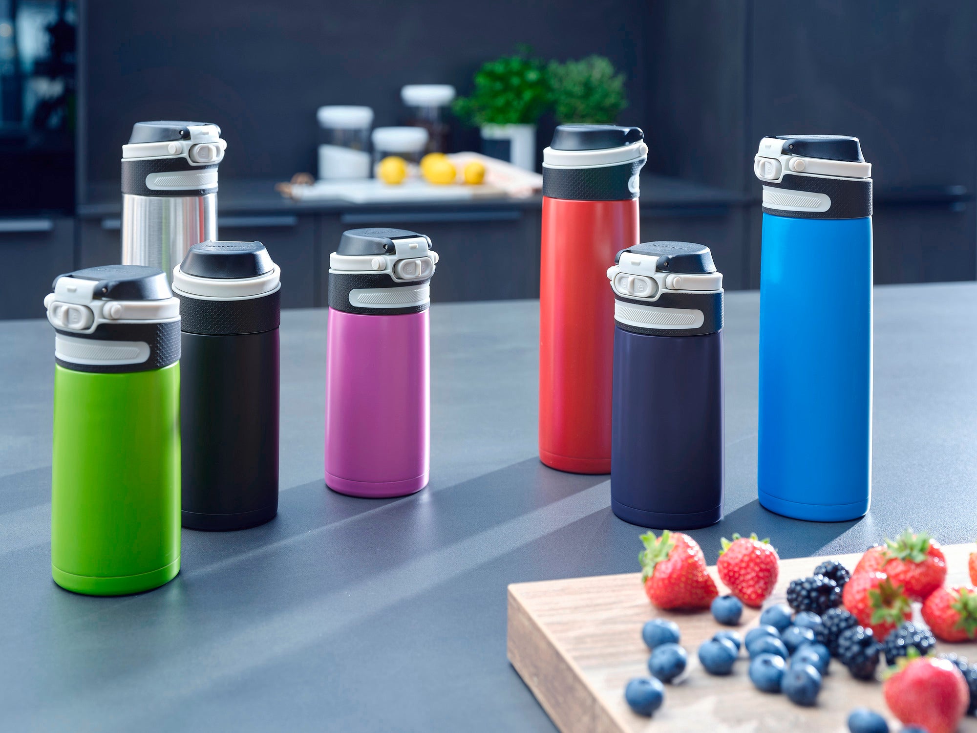 Tasses isothermes Flip de différentes couleurs et tailles sur un plan de travail de cuisine. Au premier plan, une planche en bois avec des baies fraîches. Idéal pour les déplacements.