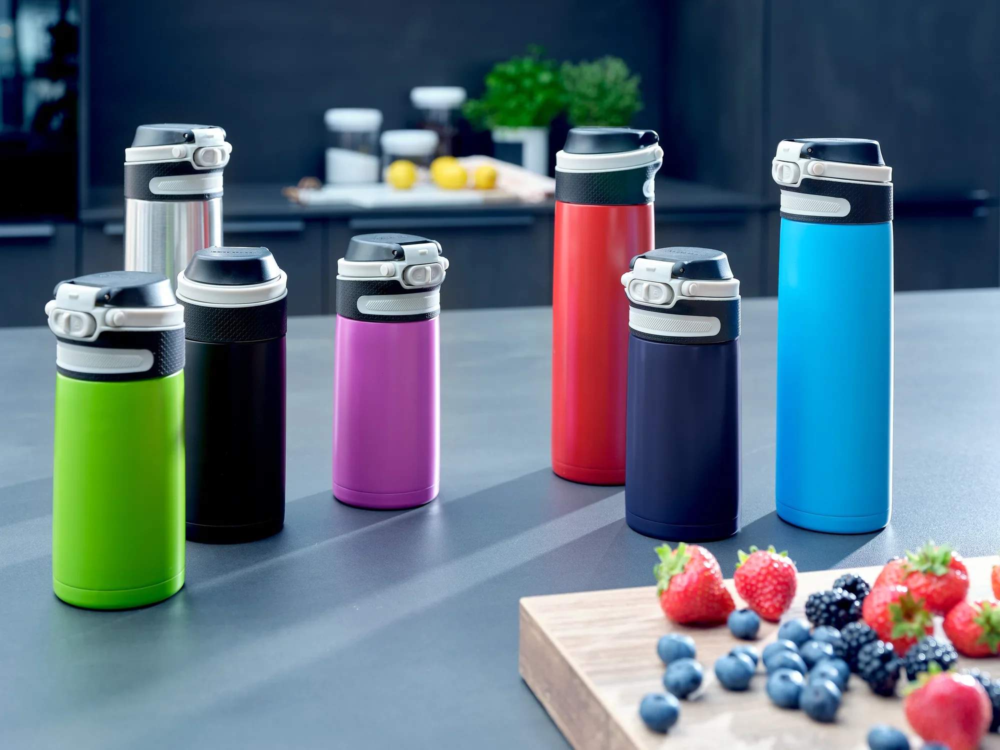 Izolowany kubek Flip w różnych kolorach i rozmiarach na blacie kuchennym. Na pierwszym planie drewniana deska ze świeżymi jagodami. Idealny w podróży.