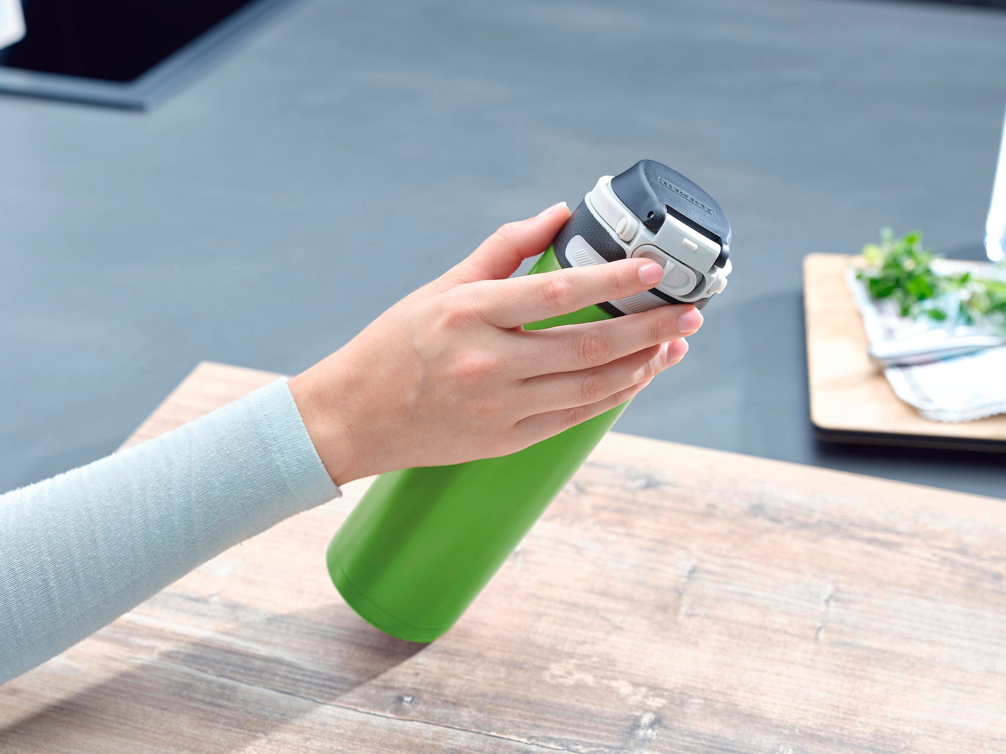 Eine Hand hält einen grünen Isolierbecher Flip mit 600 ml Fassungsvermögen. Der Becher hat einen schwarzen Deckel und liegt auf einem Holztisch in einer modernen Küche.