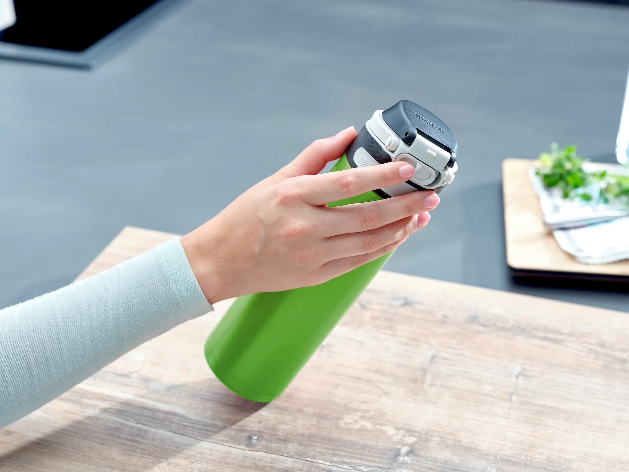 Jedna ręka trzyma zielony kubek próżniowy Flip o pojemności 600 ml. Kubek ma czarną pokrywkę i stoi na drewnianym stole w nowoczesnej kuchni.