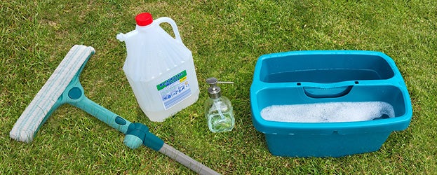 Ein Fensterwischer, ein Kanister Reiniger, eine Seifenspenderflasche und ein Eimer mit Seifenwasser stehen auf dem Rasen.