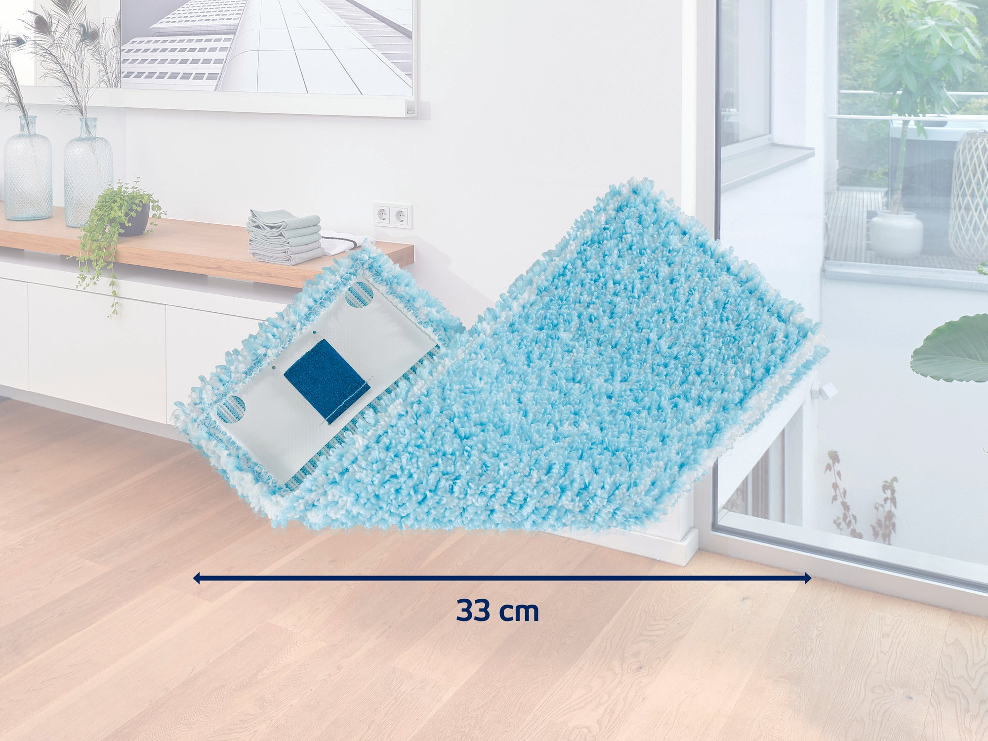 Een turquoise gekleurde mop met een breedte van 33 cm ligt op een lichtgekleurde houten vloer in een moderne kamer. Op de achtergrond zijn een raam en een witte ladenkast te zien.