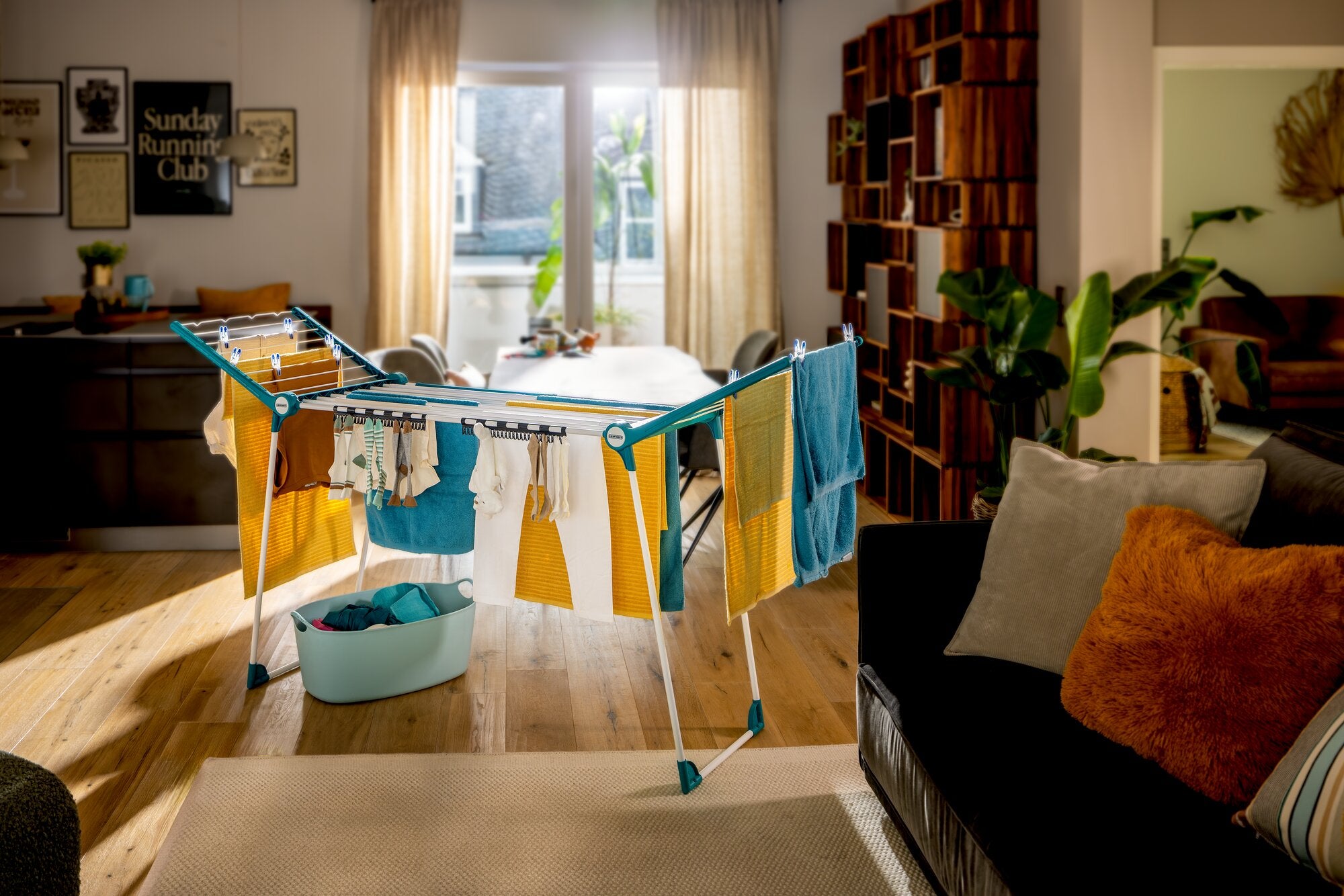 Leifheit Pegasus RockSolid Wäscheständer in einem warmen Wohnraum mit aufgehängter bunter Wäsche, seitlich ausgeklappten Flügeln und stabilem Gestell vor Fenster, Regal und Sofa.