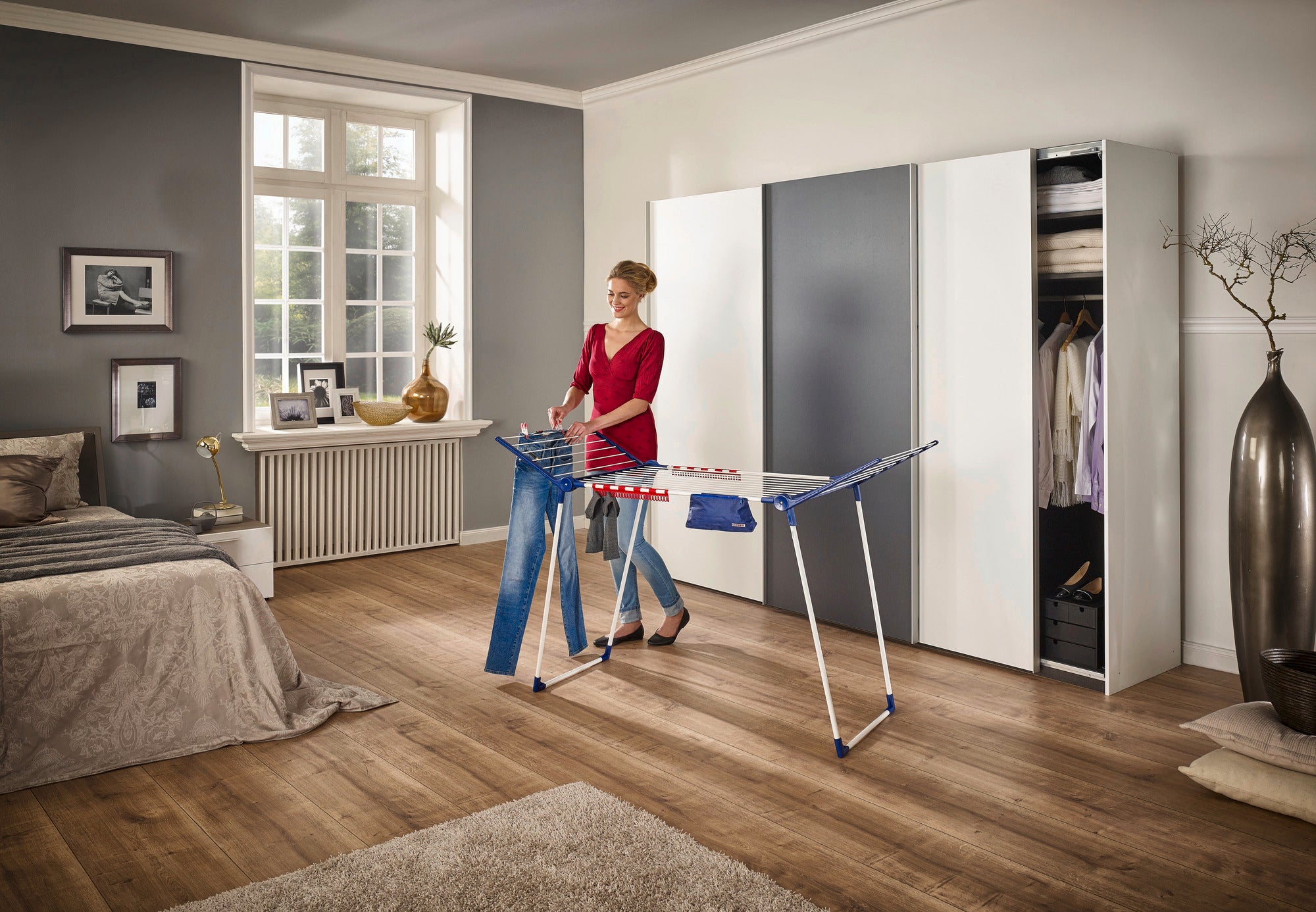 Eine Frau benutzt den Standtrockner Pegasus 180 Solid Plus in einem modernen Schlafzimmer. Der Trockner bietet viel Platz für Wäsche und ist ideal für große Räume.