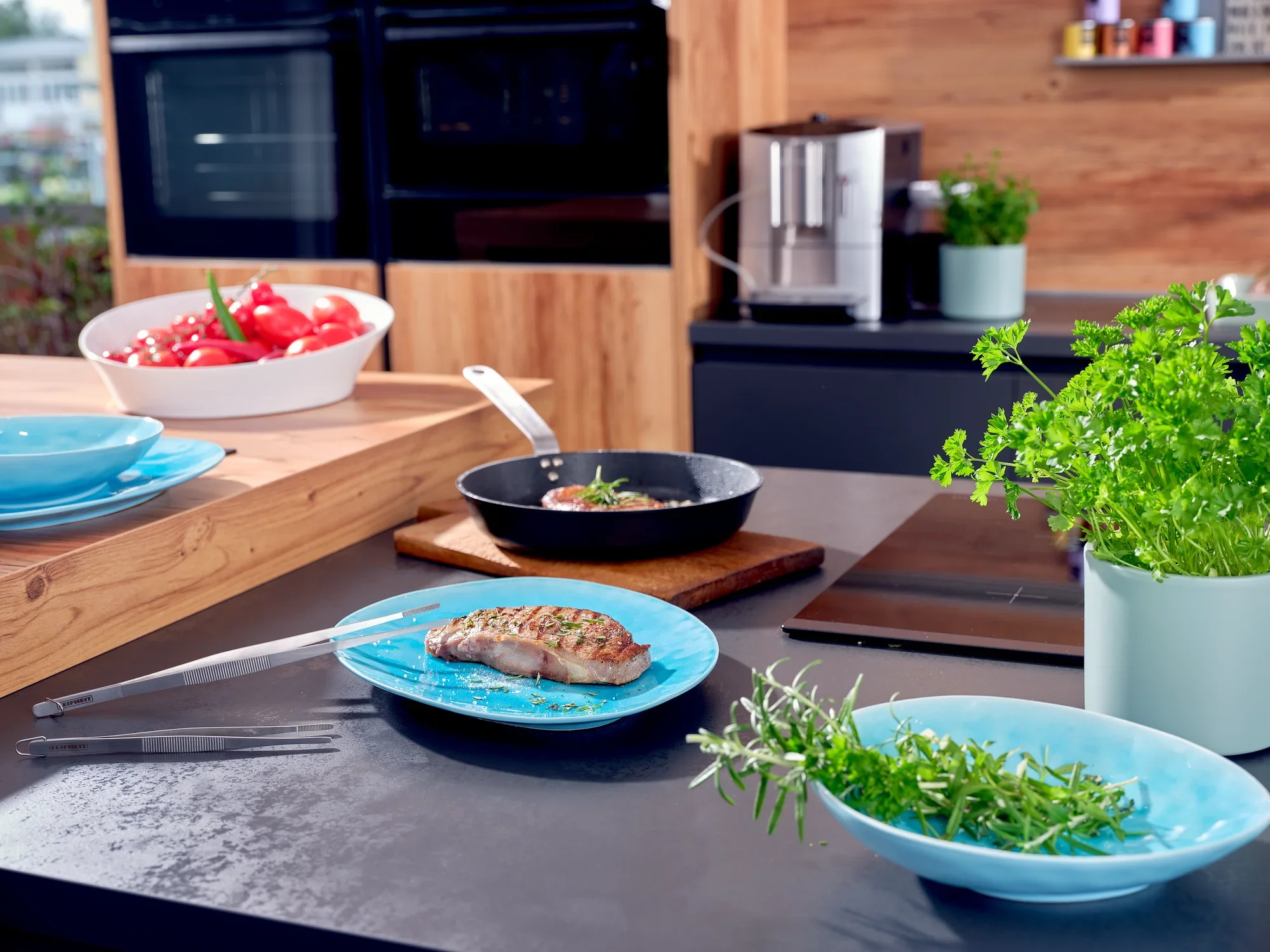 In een moderne keuken ligt een 20 cm lange keukenpincet naast een bord gebraden vlees en verse kruiden. Op de achtergrond zie je tomaten en een koffiezetapparaat.