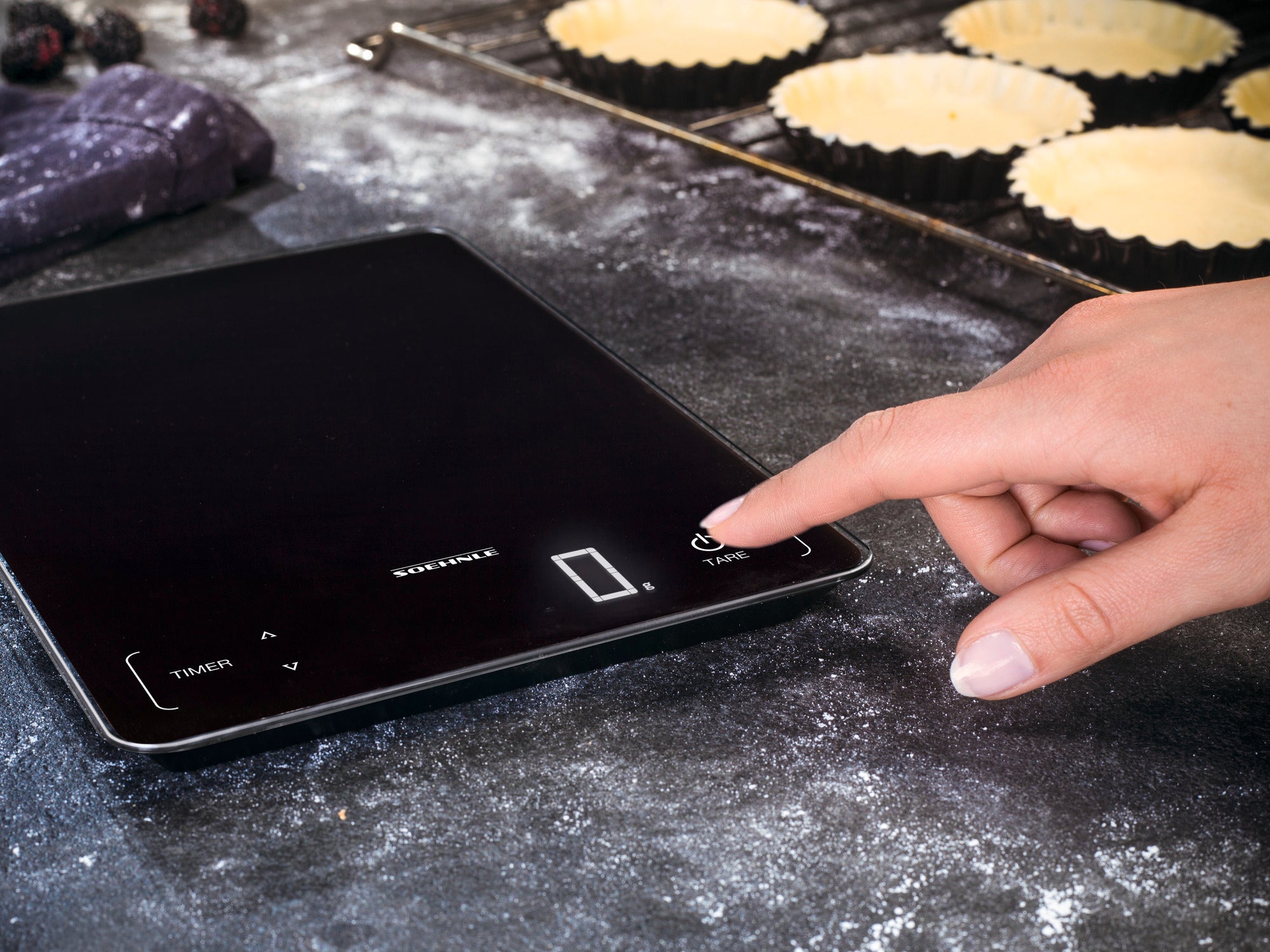 A hand operates the Page Profi 100 digital kitchen scale on a work surface. Baking tins and flour can be seen in the background, suggesting that someone is preparing to bake.